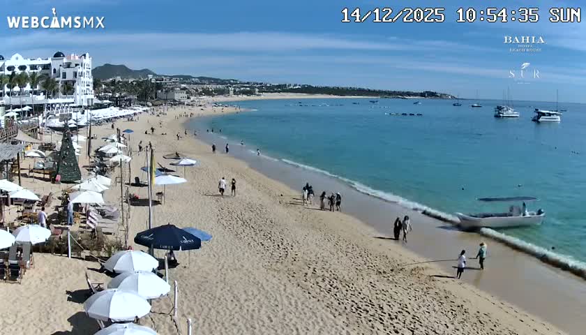 A serene night scene captures a resort beach with tables and string lights on the sand, gentle waves meeting the shore, and several boats anchored in the calm ocean under a clear, dark sky, with distant city lights illuminating the horizon.