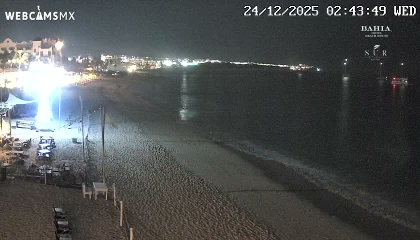 A serene night scene captures a resort beach with tables and string lights on the sand, gentle waves meeting the shore, and several boats anchored in the calm ocean under a clear, dark sky, with distant city lights illuminating the horizon.