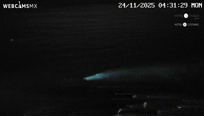 A prominent, glowing turquoise-blue trail stretches horizontally across a dark night scene, with faint, textured reflections visible on the dark surface below, under clear conditions.