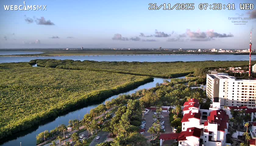 An elevated nighttime view reveals a cloudy sky over a dark body of water reflecting a distant illuminated city skyline, with brightly lit hotels, streets, and palm trees in the foreground.