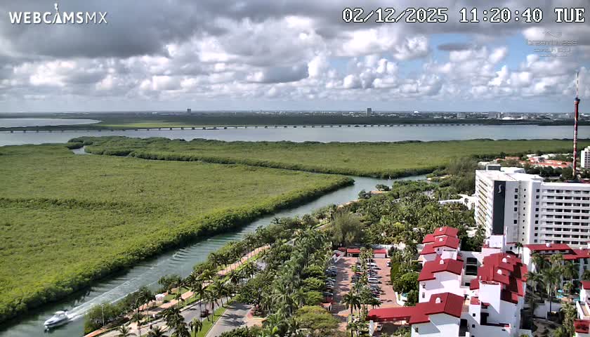 An elevated view captures a tropical scene featuring a winding canal with a boat, lush green vegetation, resort buildings, a vast lagoon with a bridge, and a distant city skyline under a partly cloudy sky.