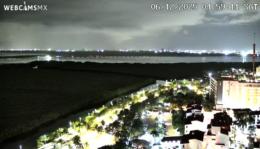 An aerial night view reveals a vibrant coastal city with brightly lit buildings, palm-lined streets, and a canal leading to a large body of water reflecting distant city lights, all under a heavily cloudy sky.