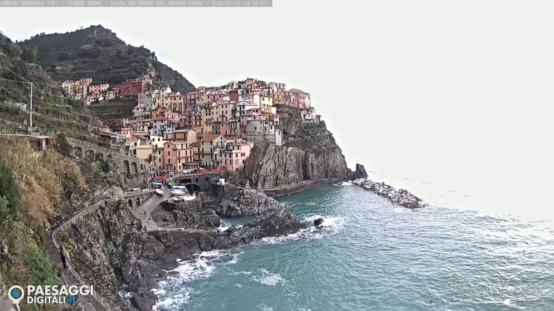 A colorful village clings to a steep, rocky hillside overlooking a partially calm sea under an overcast sky.