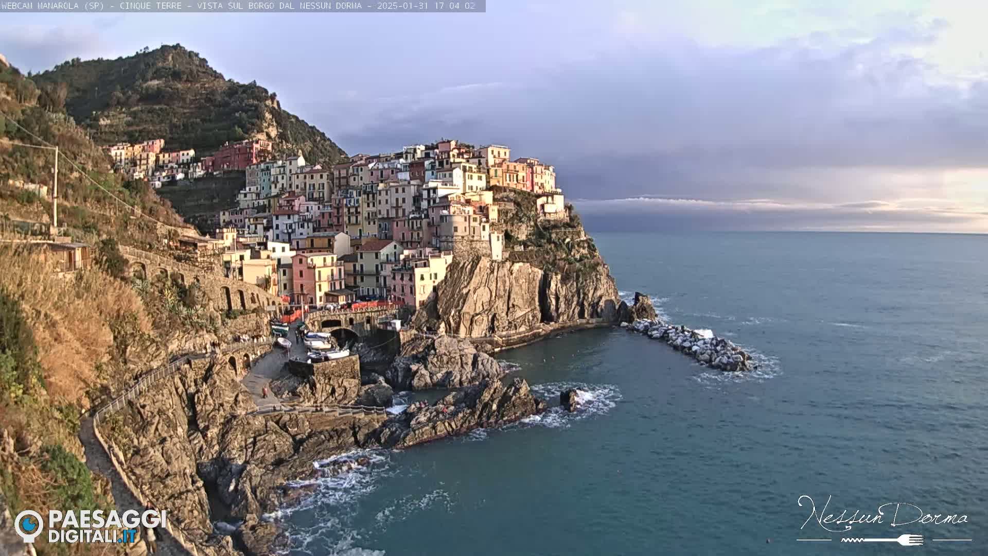 A colorful village clings to a rocky cliffside overlooking a calm sea under a partly cloudy sky.