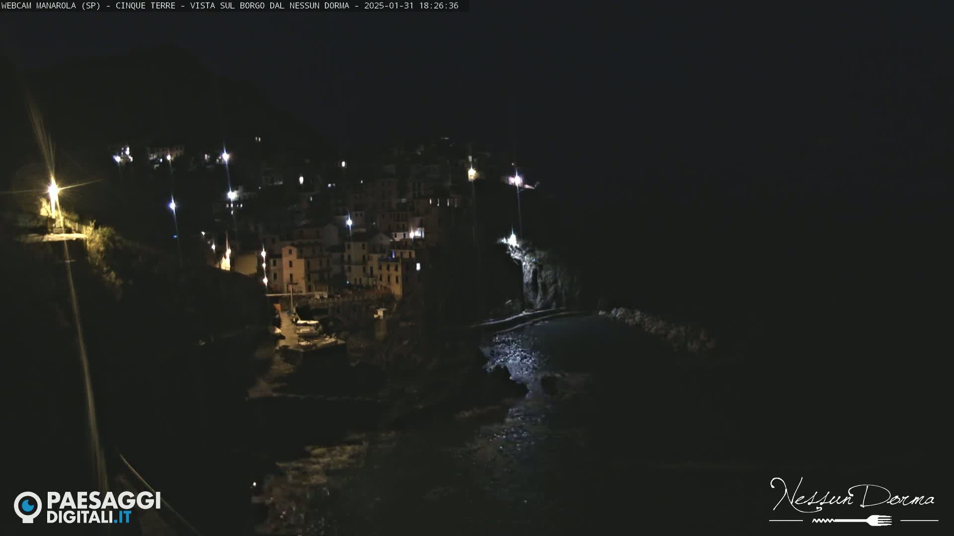 A nighttime view of a coastal village illuminated by scattered lights, with dark water visible below.
