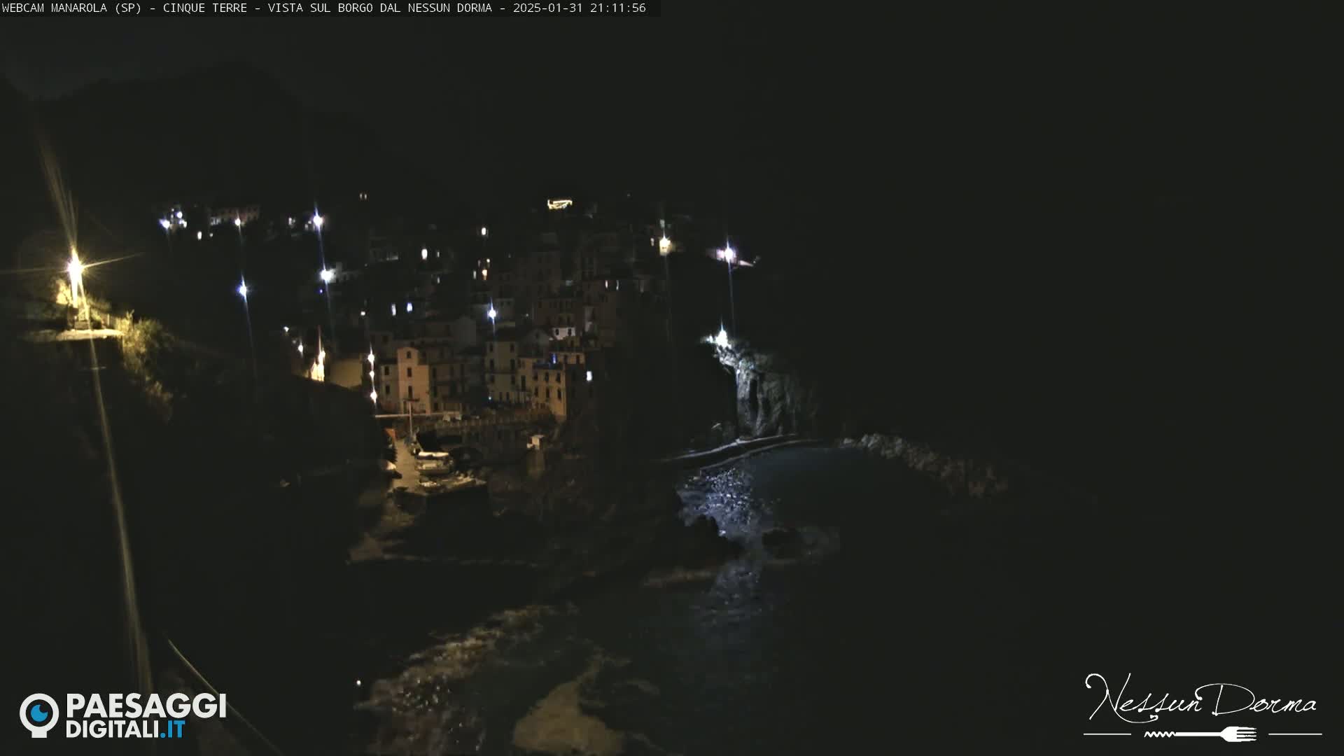 A nighttime, aerial view of a coastal town with dimly lit buildings and a dark, rocky shoreline.
