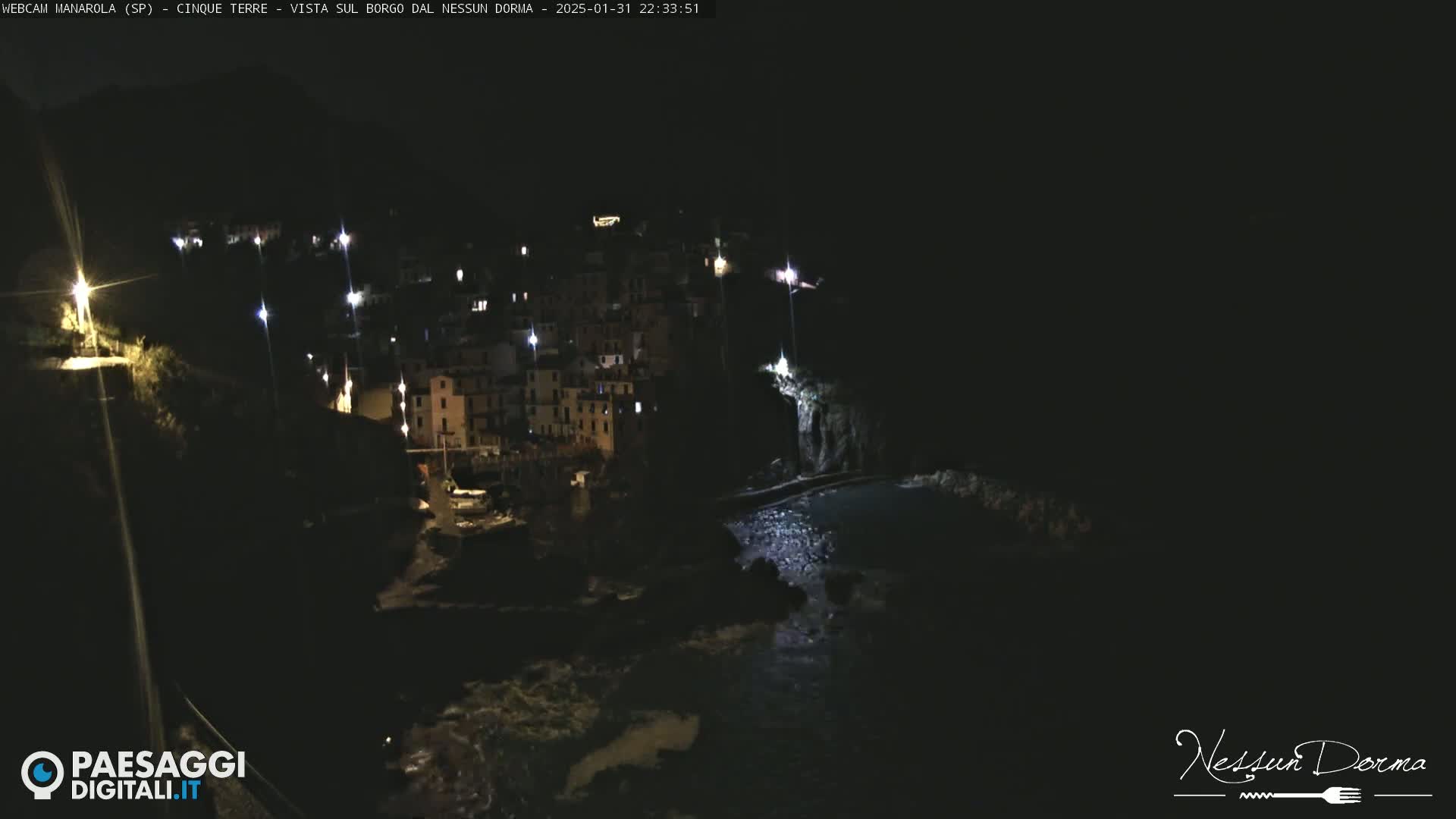 A nighttime, coastal village with dimly lit buildings is seen from above, next to dark water.