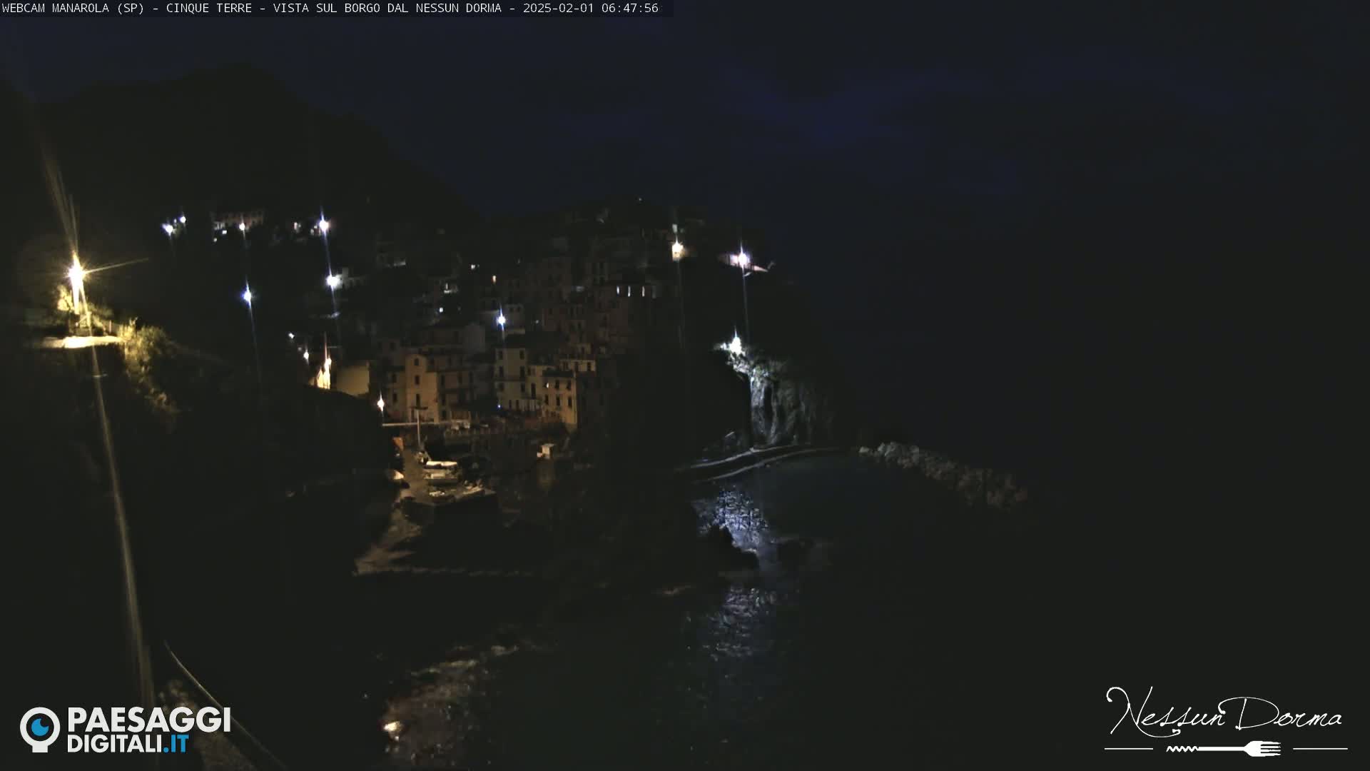 A nighttime view of a coastal village illuminated by scattered lights, with dark water below.