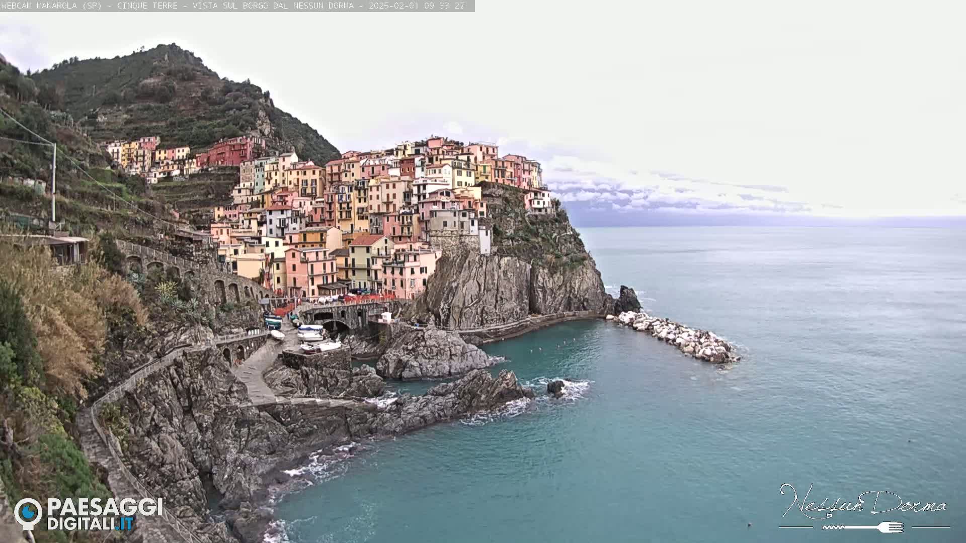 A colorful village clings to a steep cliffside overlooking a calm, teal sea under a mostly cloudy sky.