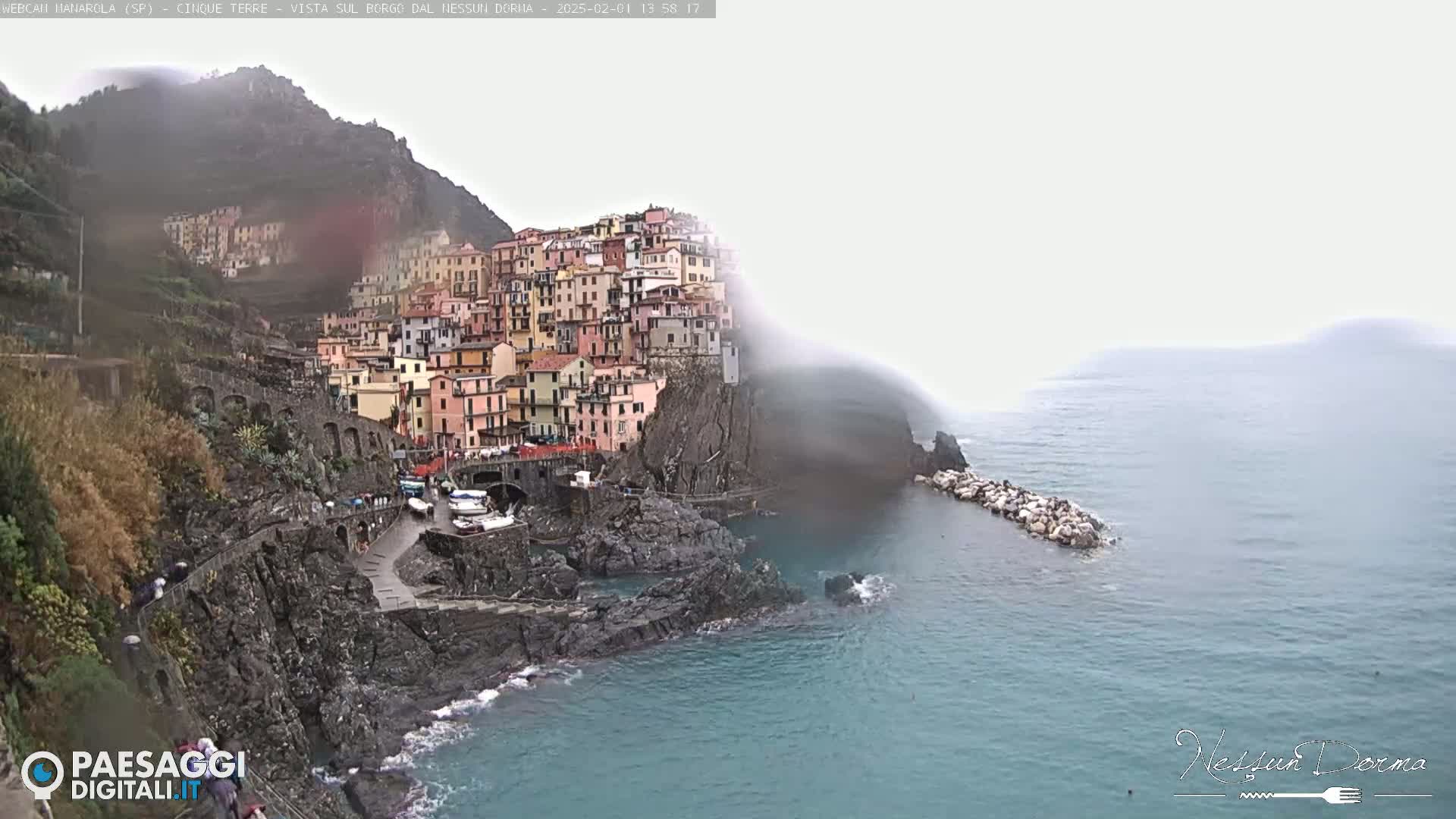 A colorful village clings to a steep cliffside overlooking a calm, foggy sea.