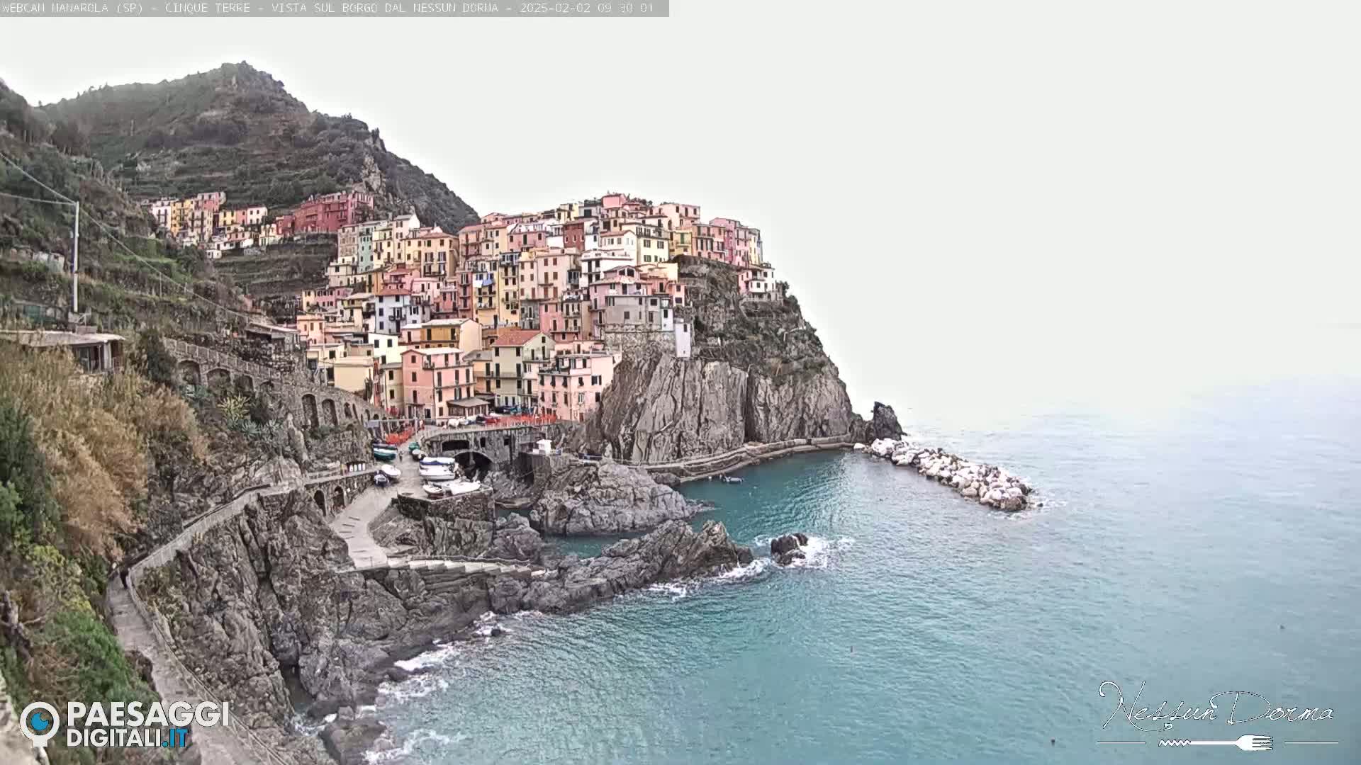 A colorful village clings to a rocky cliffside overlooking a calm sea under an overcast sky.