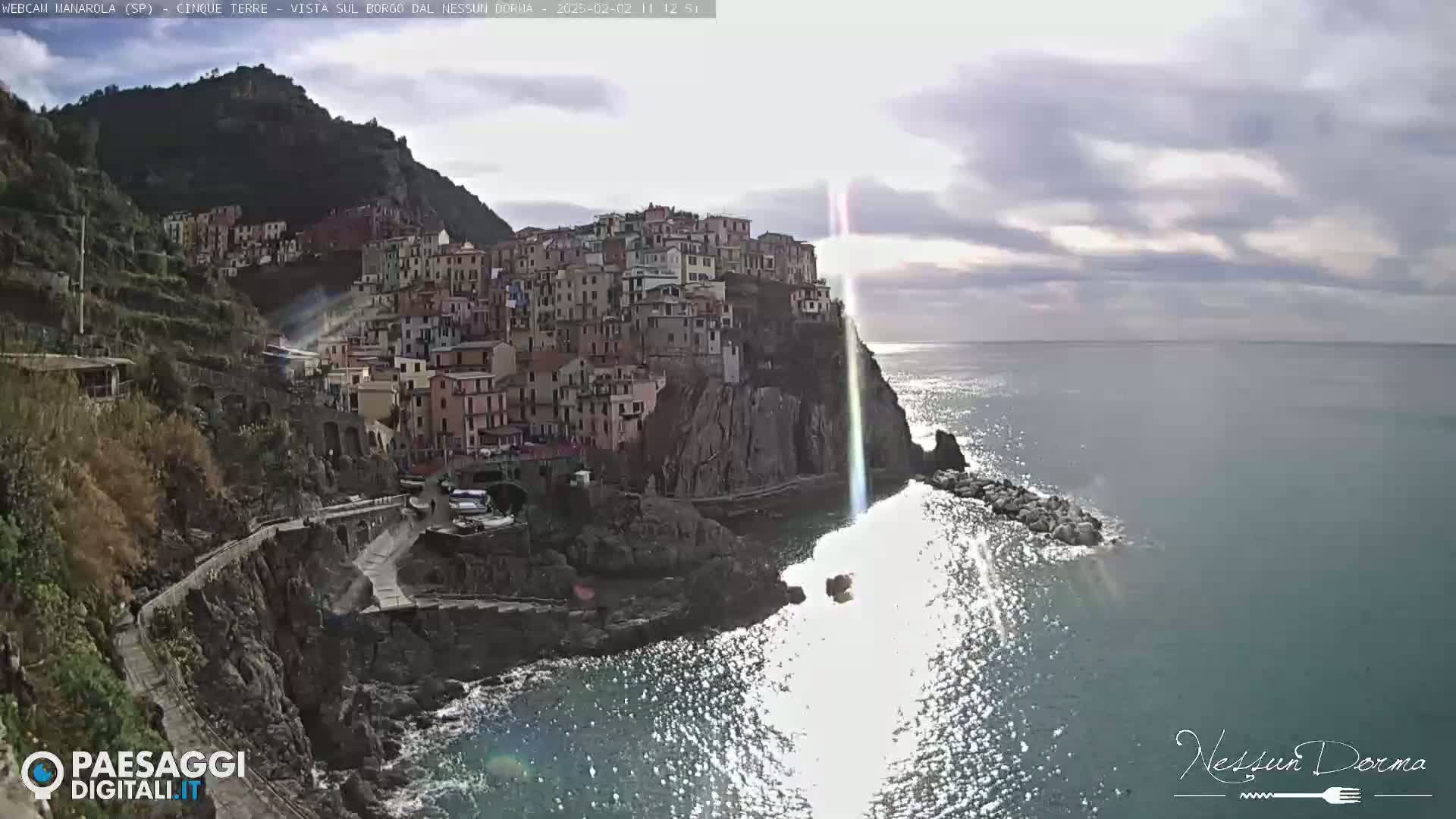 A sunlit coastal village clings to a cliffside overlooking a sparkling sea under a partly cloudy sky.