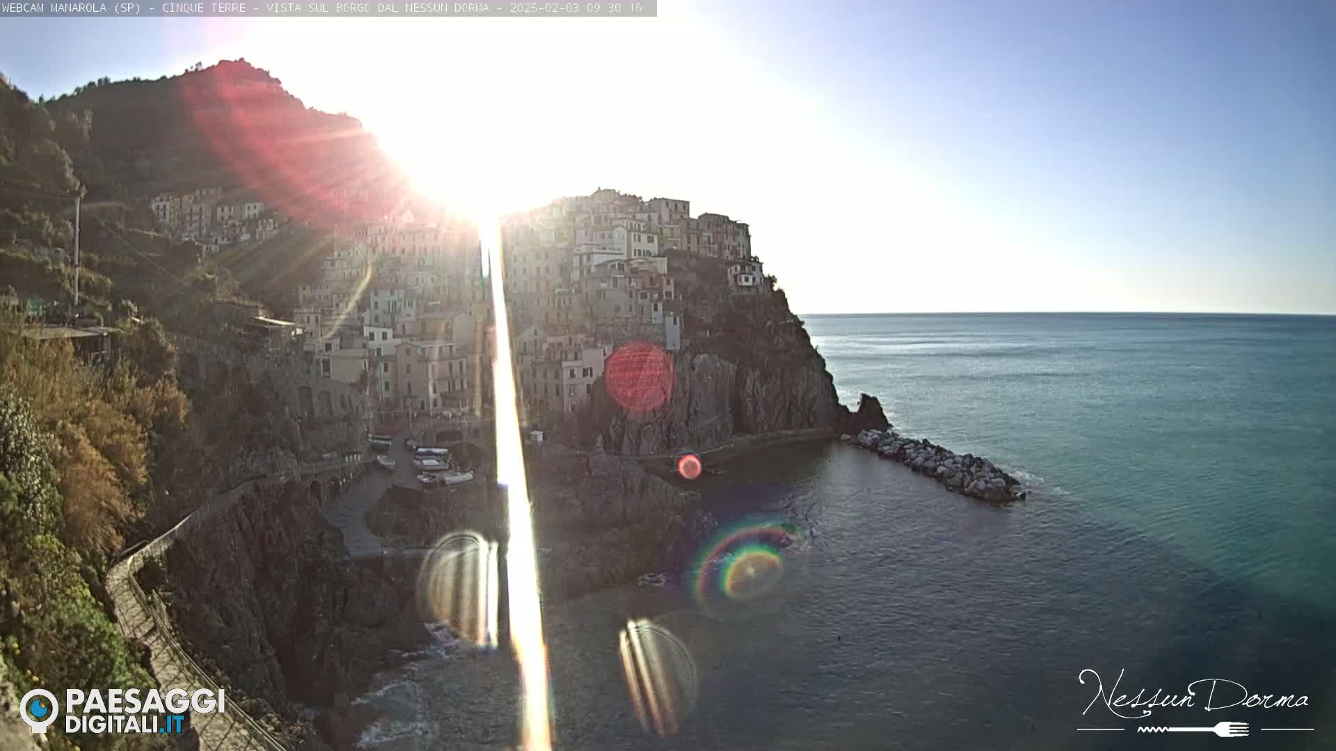 A sunlit coastal village clings to a rocky cliffside overlooking a calm sea.