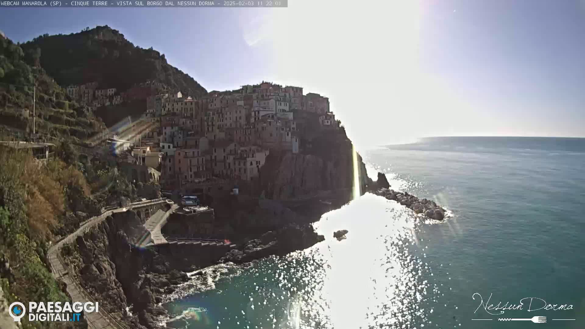 A brightly lit coastal town clings to a steep cliffside overlooking a sparkling blue sea under a mostly sunny sky.