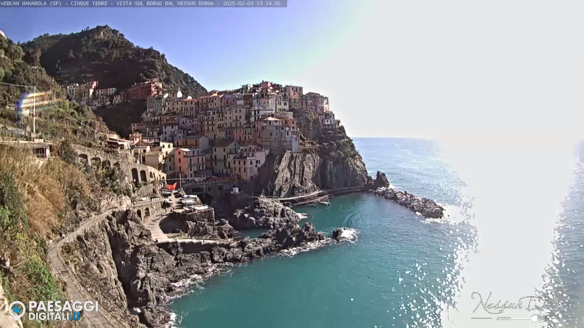 A colorful village clings to a steep cliffside overlooking a calm, blue sea under a bright, sunny sky.
