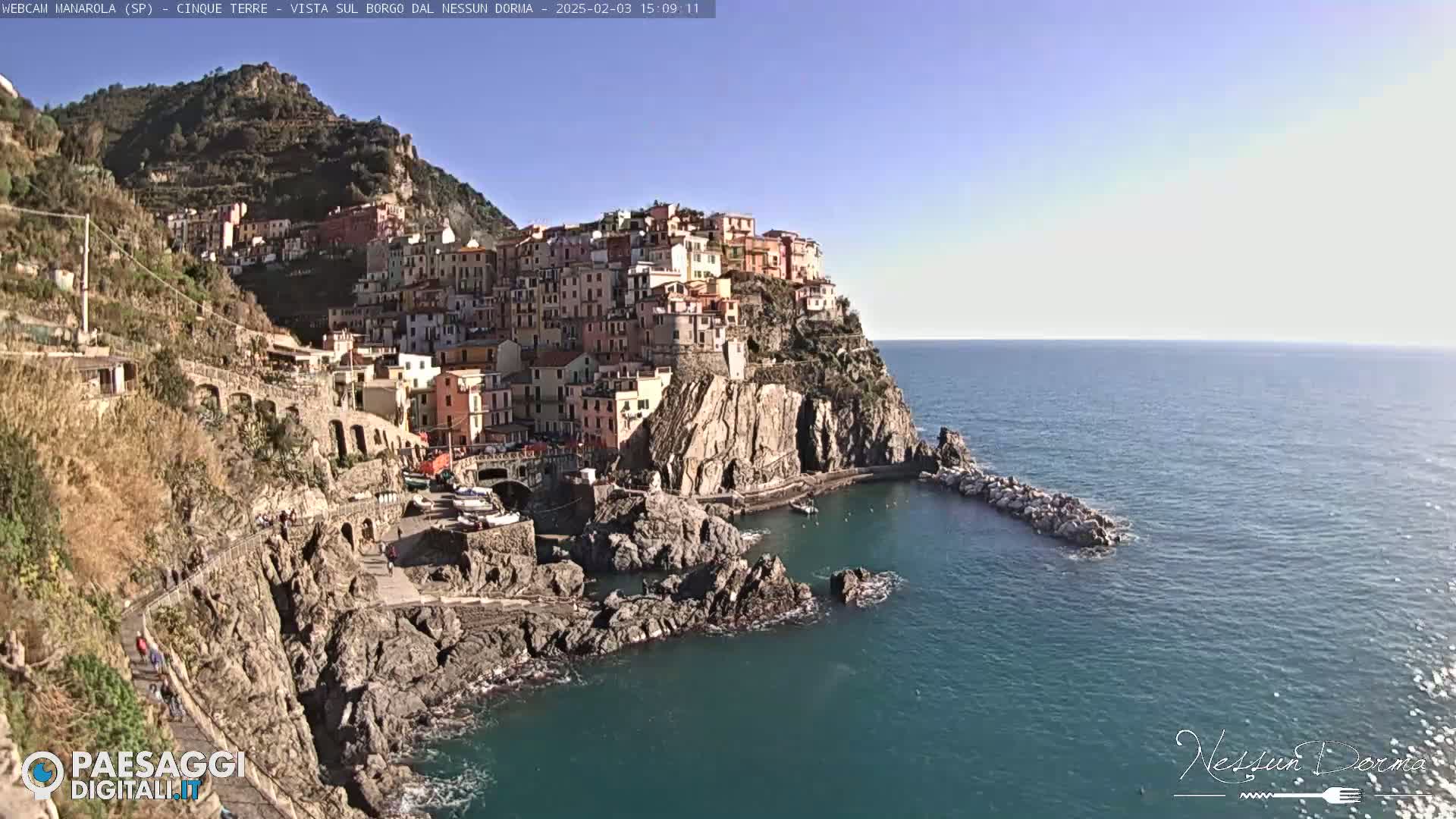 A colorful village clings to a rocky cliffside overlooking a calm, blue sea under a clear sky.