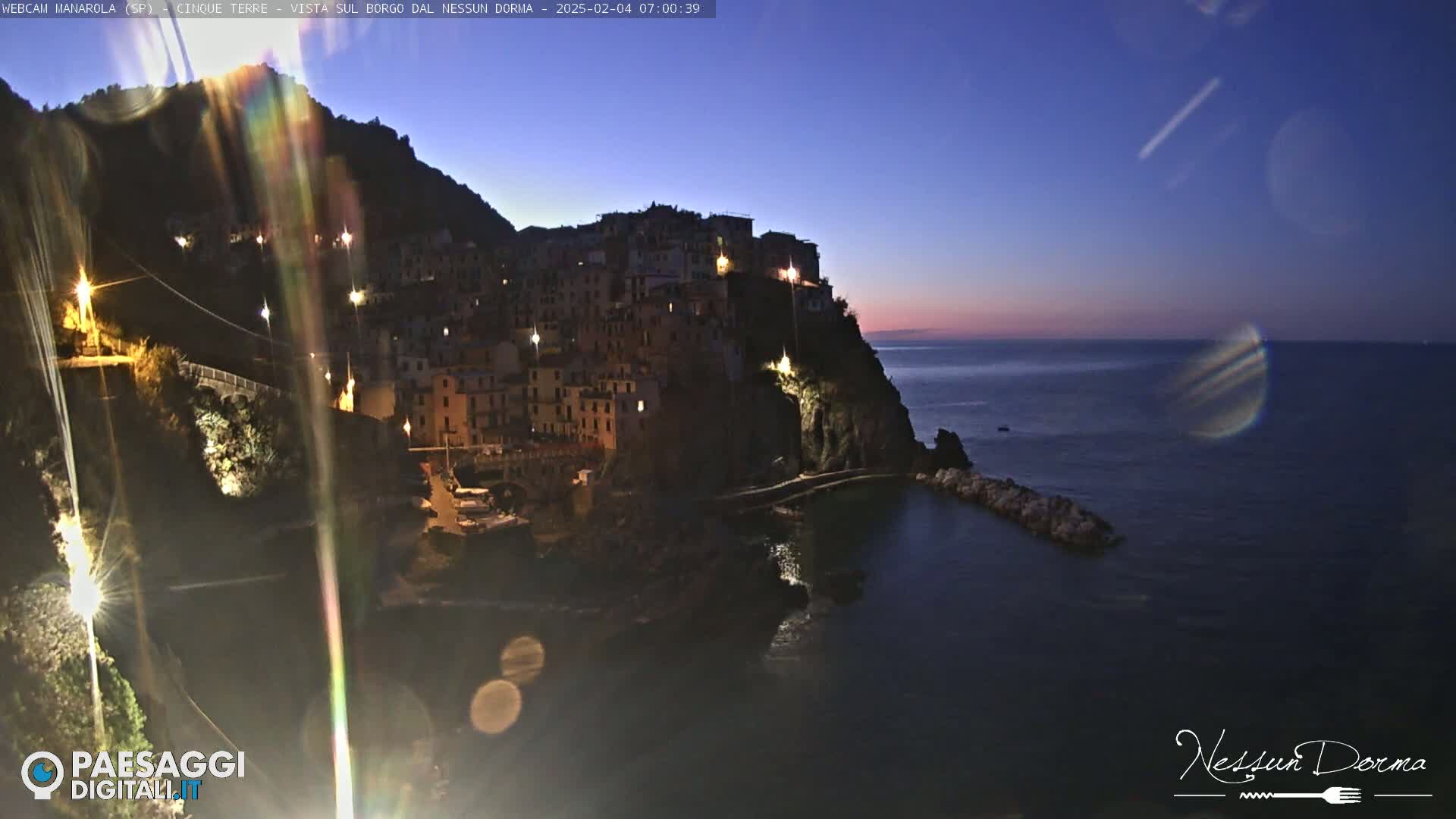 A nighttime coastal village clinging to a cliffside overlooks a dark, calm sea under a twilight sky.