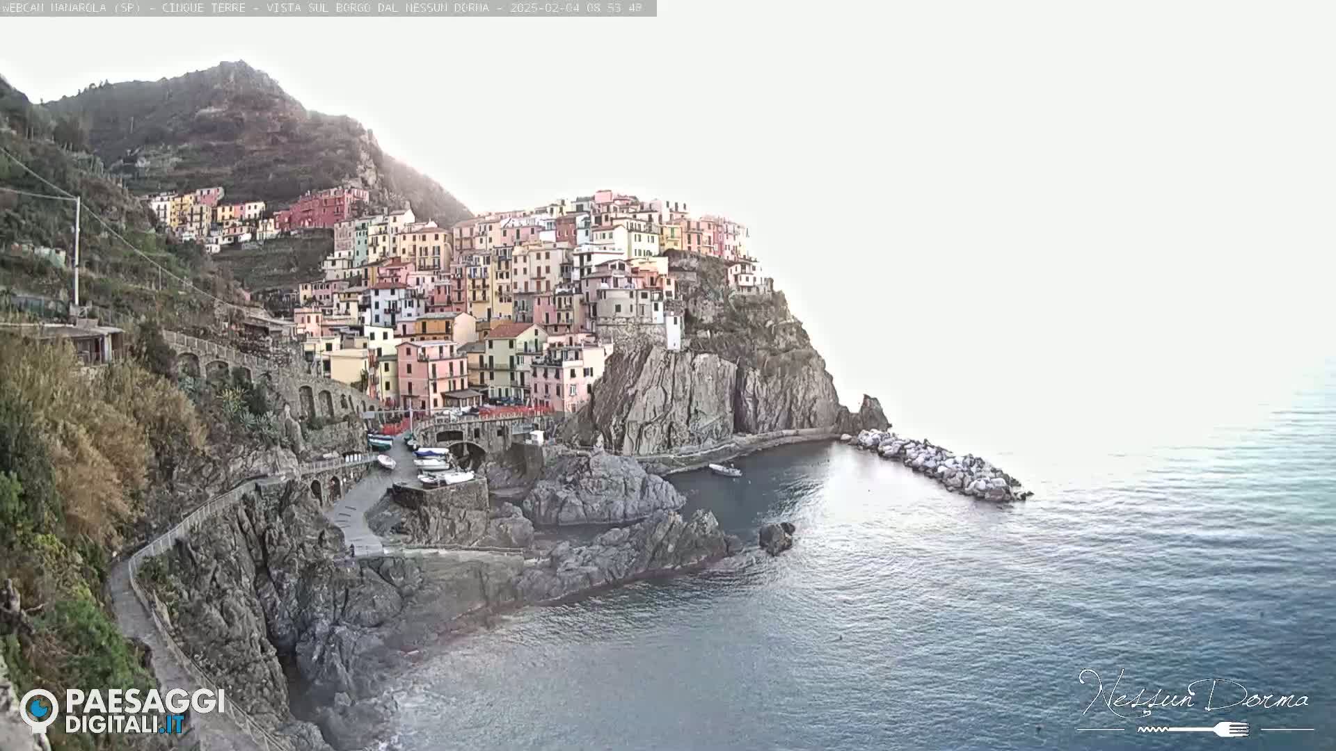 A colorful village clings to a steep cliffside overlooking a calm sea under a bright, hazy sky.