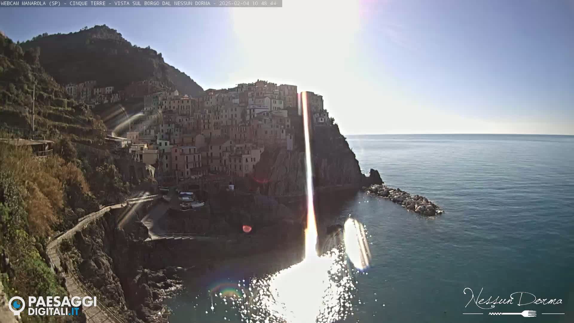 A brightly lit coastal town built into a steep cliffside overlooking a calm sea on a sunny day.