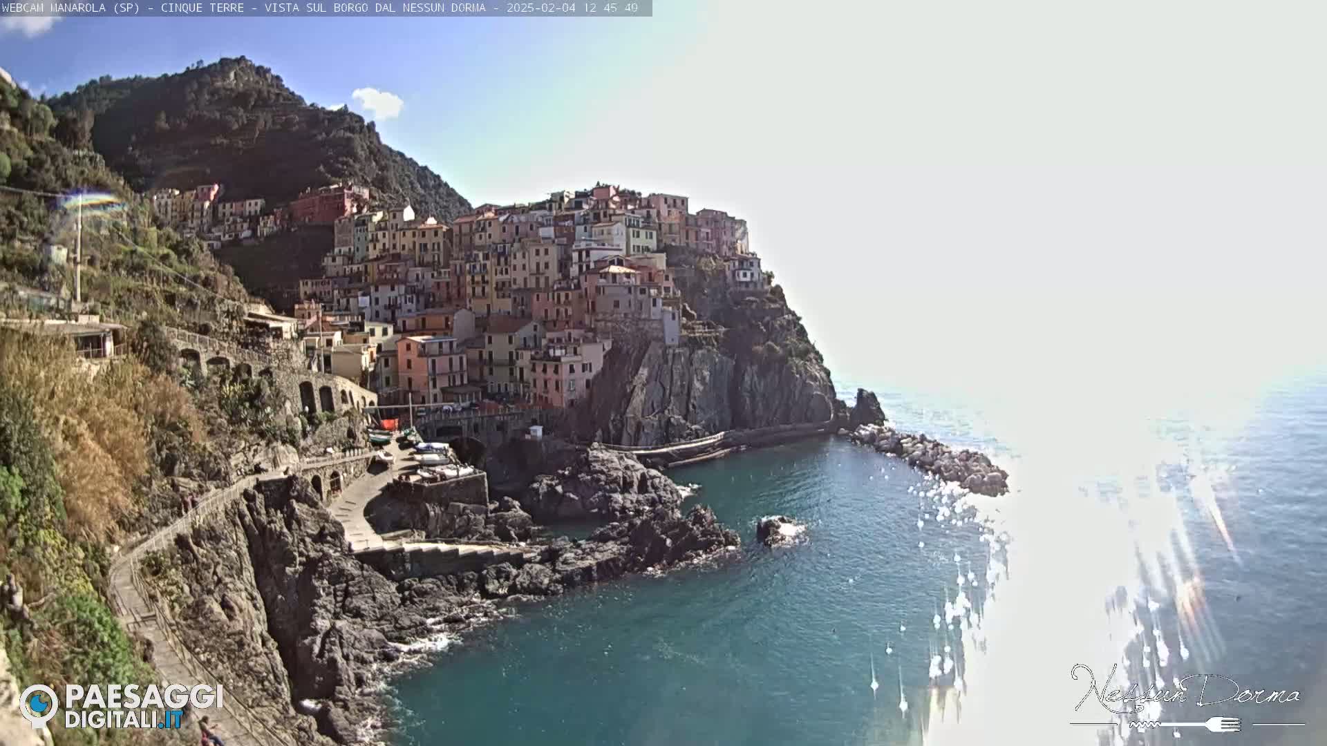 A colorful village clings to a steep cliffside overlooking a calm, blue sea under a mostly sunny sky.