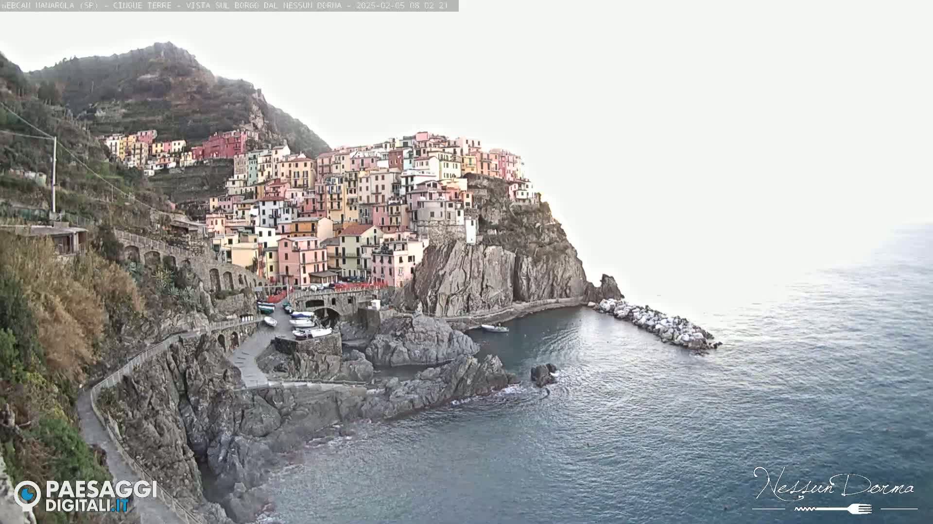 A colorful village clings to a steep cliffside overlooking a calm sea under a mostly cloudy sky.