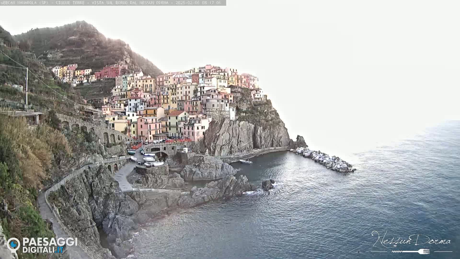 A colorful village clings to a steep cliffside overlooking a calm sea on an overcast day.