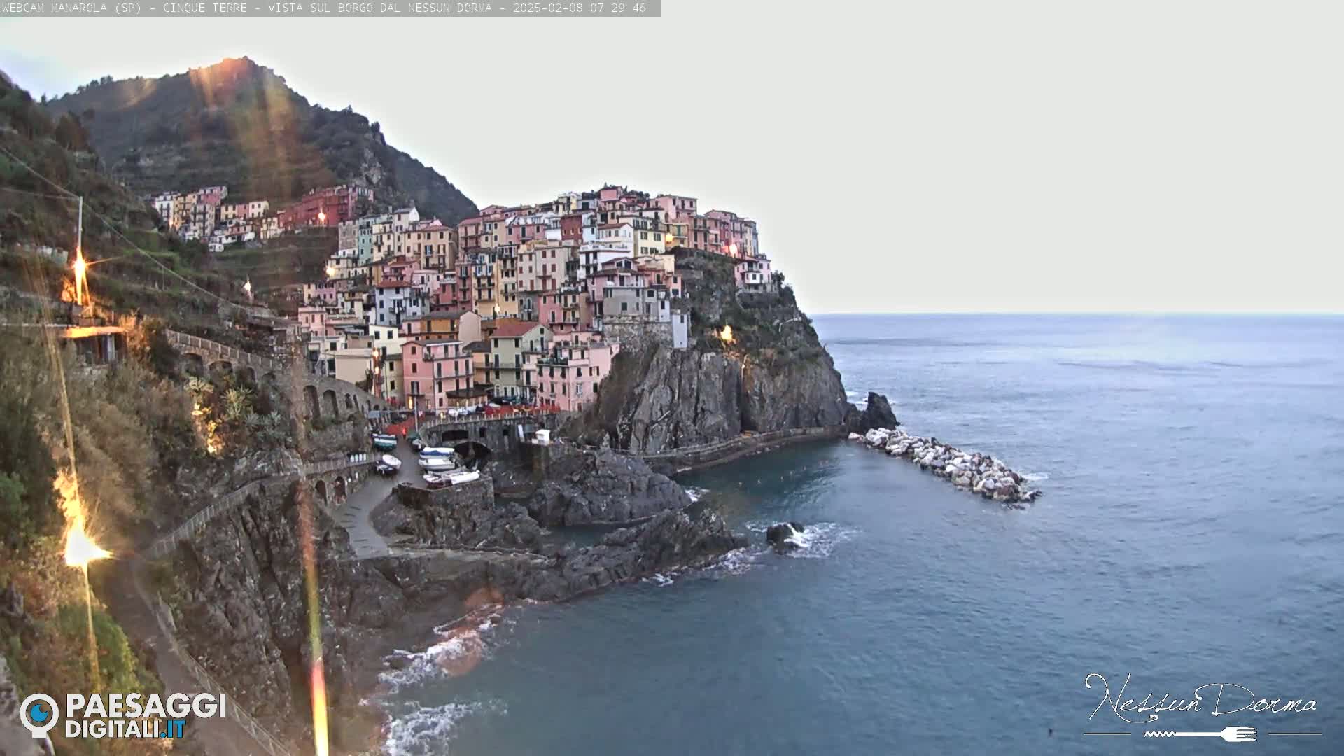 A colorful village clings to a rocky cliffside overlooking a calm sea under a bright, overcast sky.