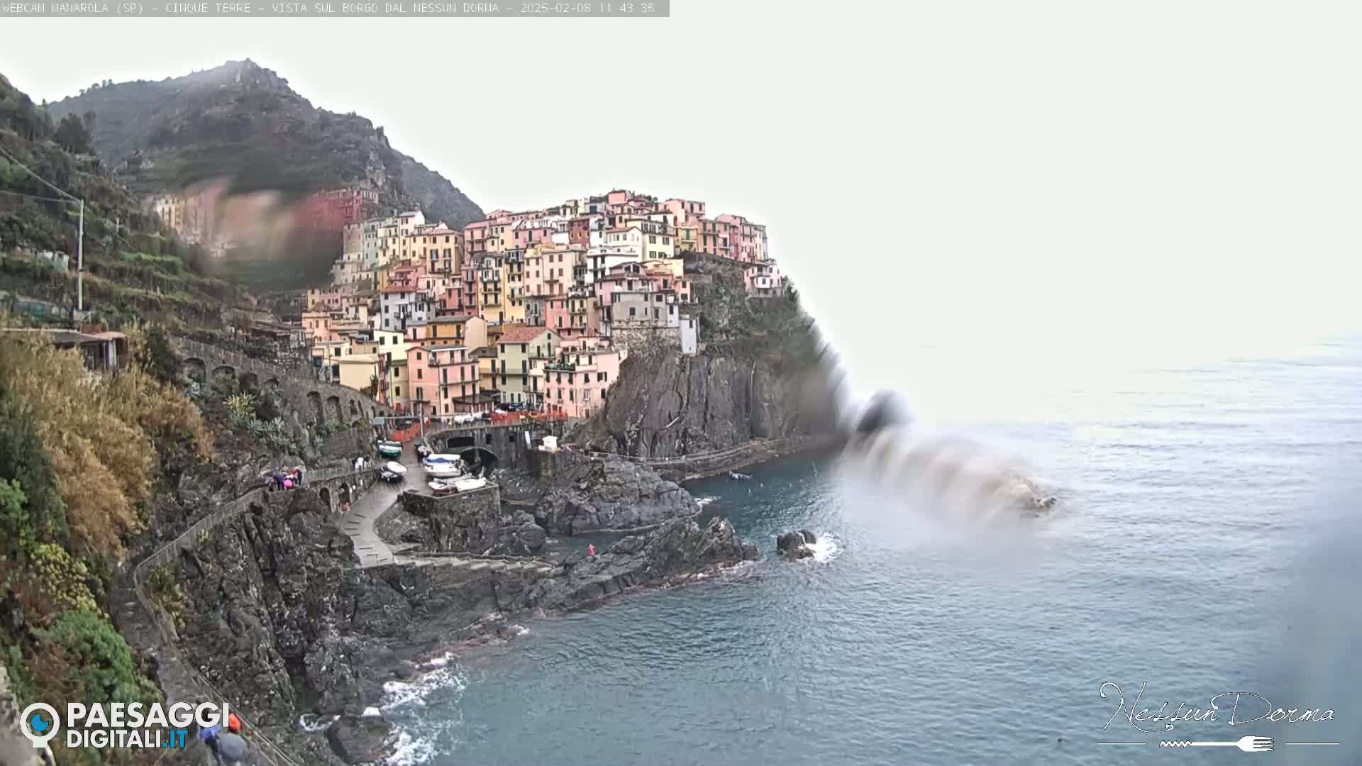 A colorful village clings to a steep cliffside overlooking a choppy sea under an overcast sky.