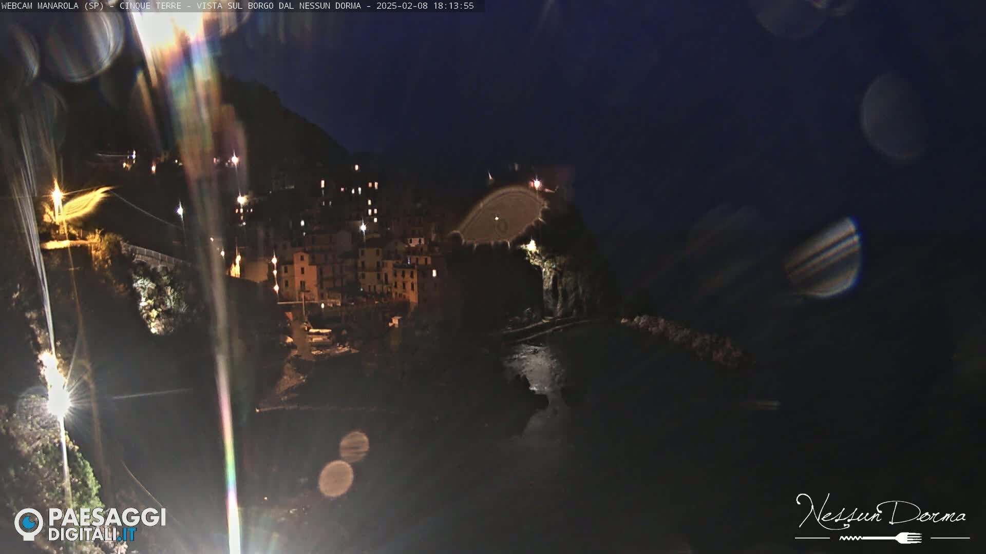 A nighttime view of a coastal town illuminated by lights, with a dark, calm body of water below and rain obscuring the view.
