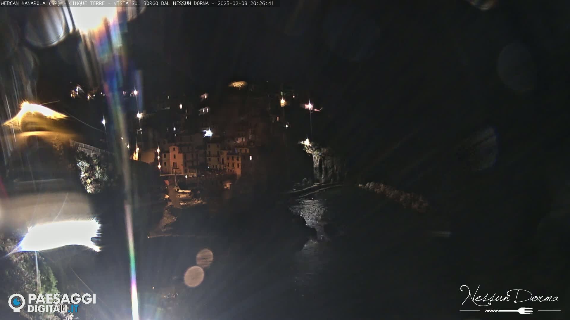 A nighttime view of a coastal town illuminated by scattered lights, with dark water visible below.