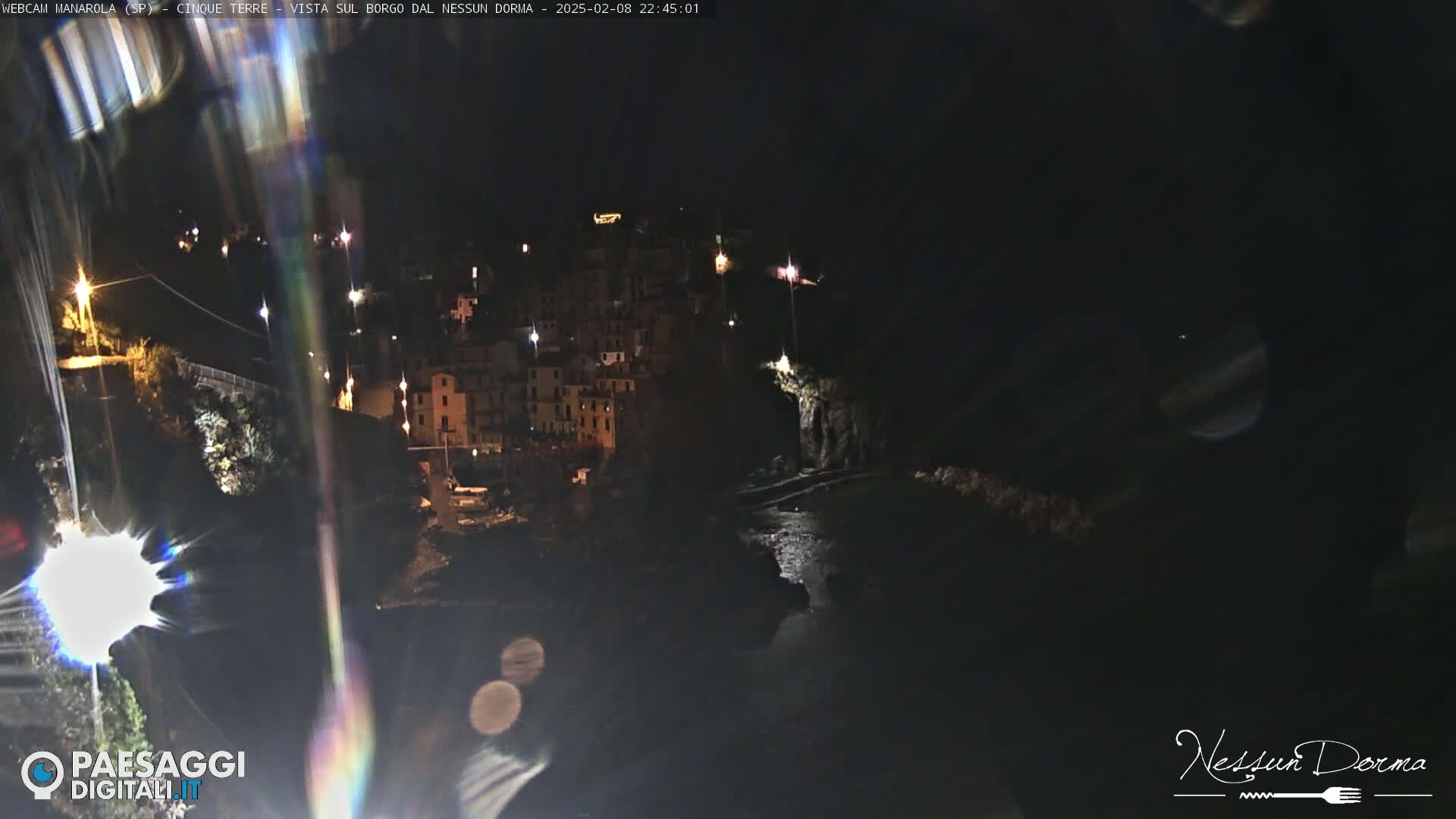 A nighttime, long shot of a coastal town illuminated by lights, with dark water visible below.