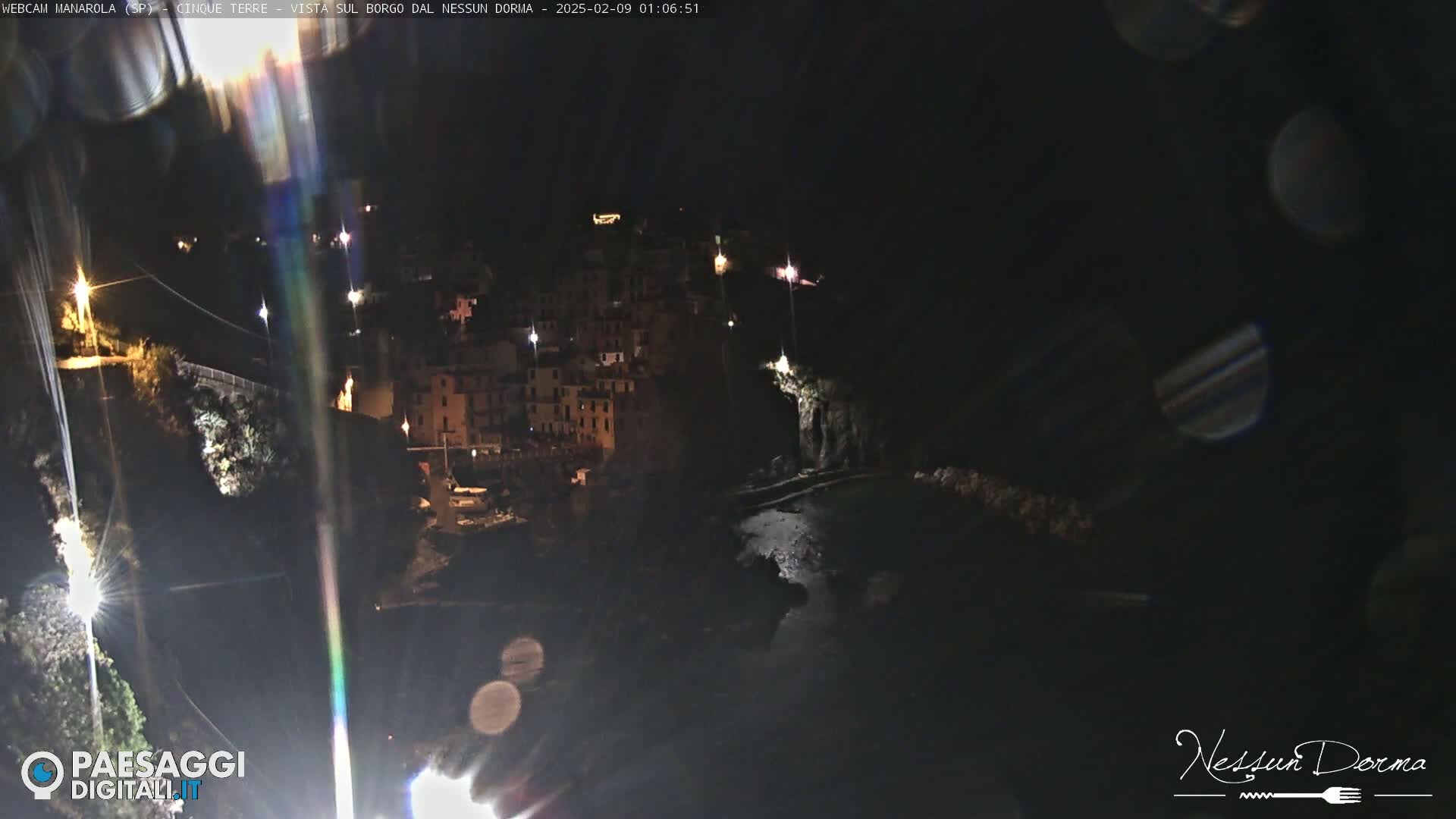 A nighttime, dimly lit coastal town is nestled against a dark hillside, partially visible from across a body of water.