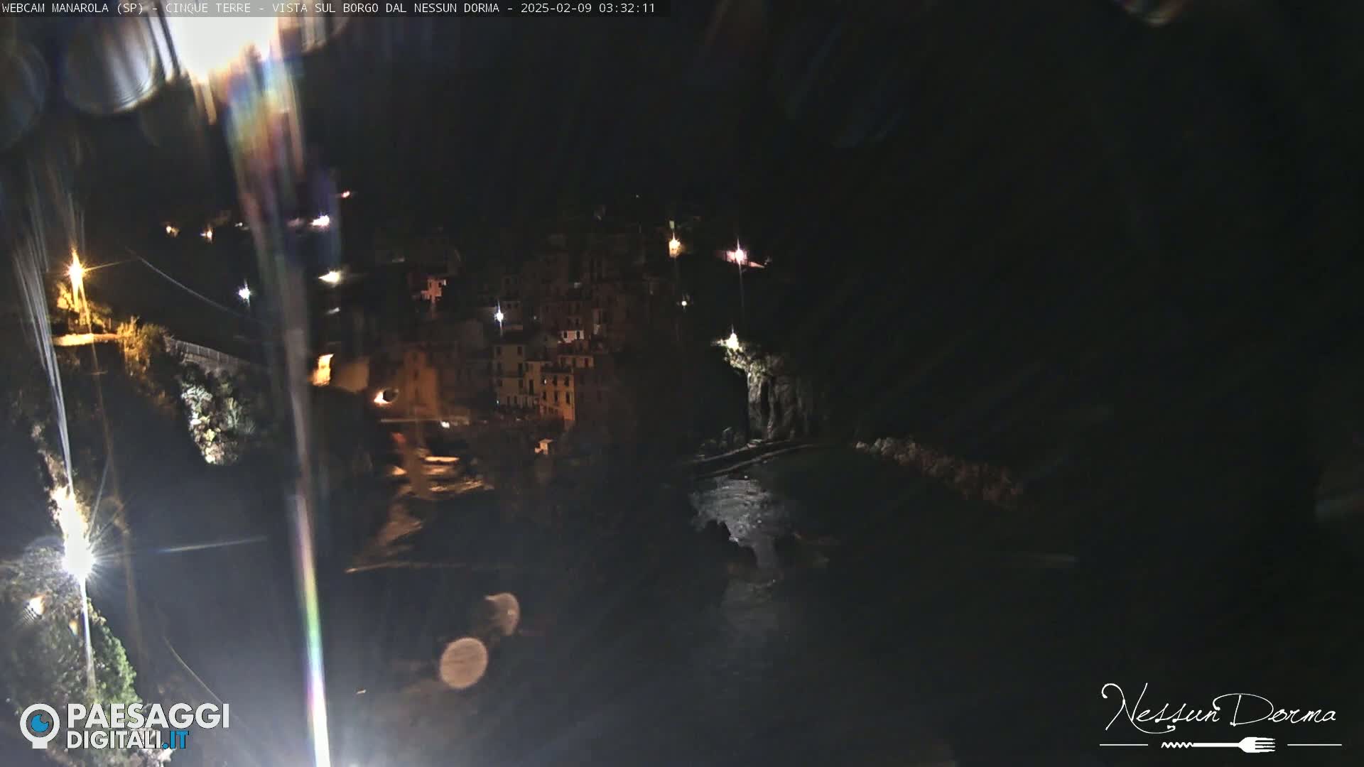 A nighttime, long shot of a coastal village with buildings lit up along a dark, rocky shoreline.