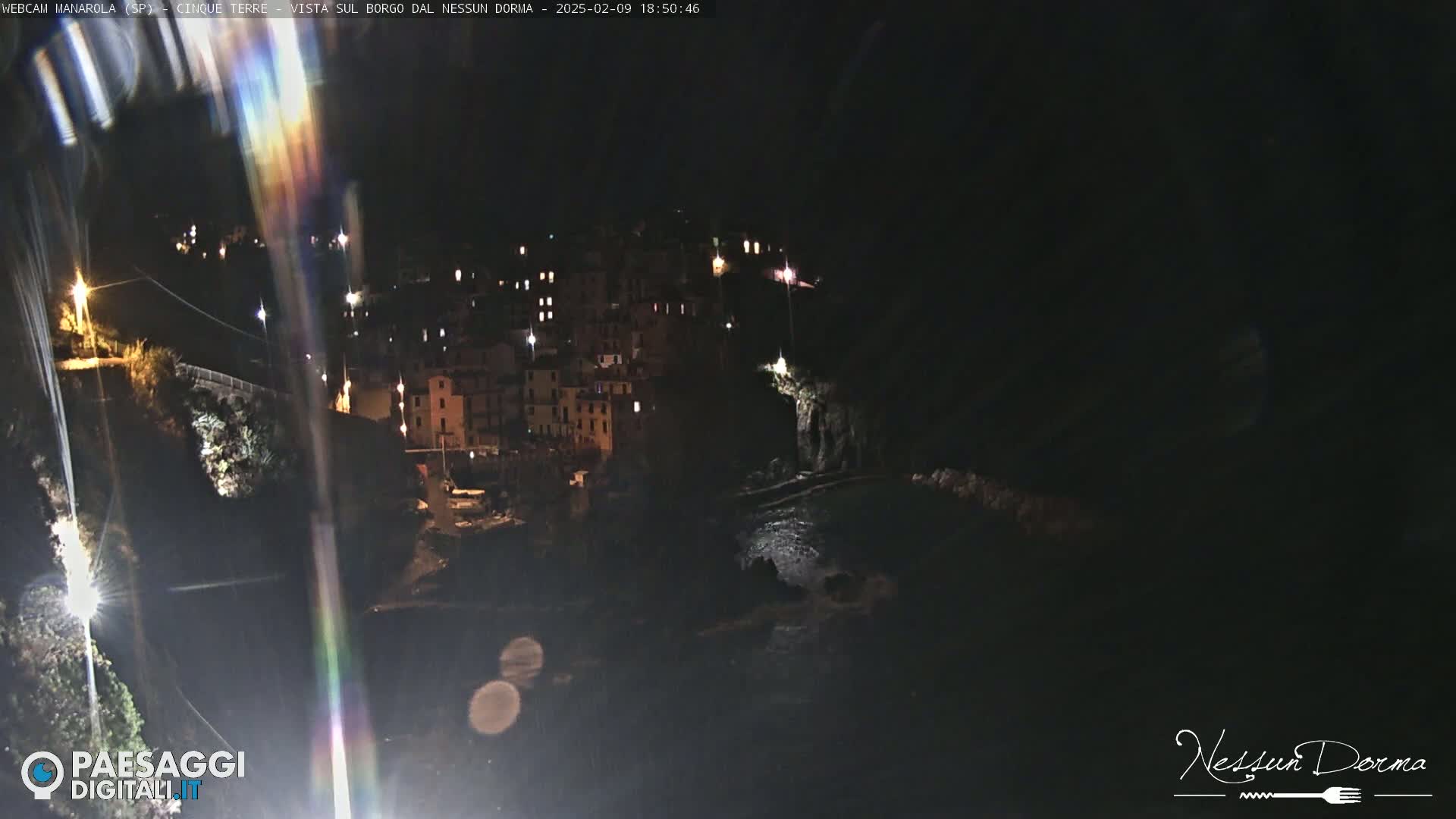A nighttime view of a coastal town illuminated by lights, with dark water visible below.
