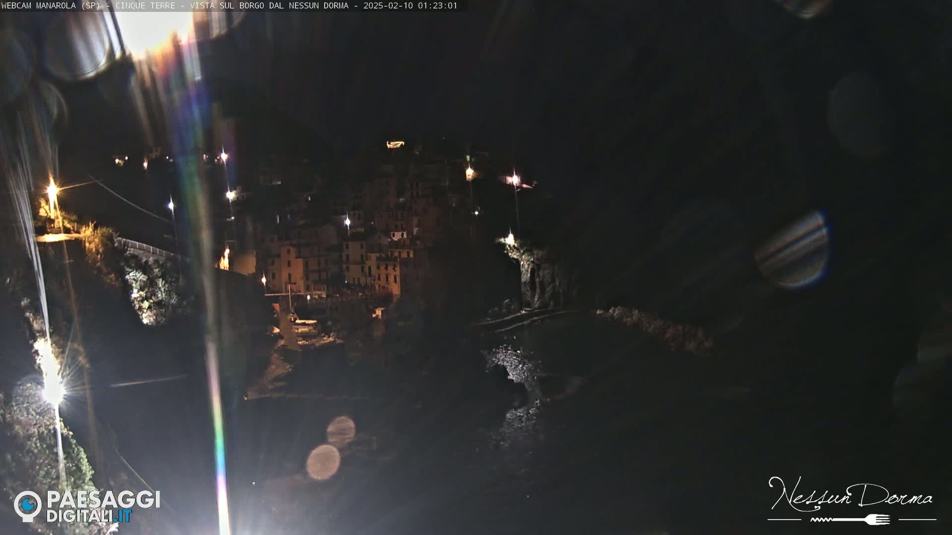 A nighttime view of a coastal town, illuminated by scattered lights, with a dark body of water below.