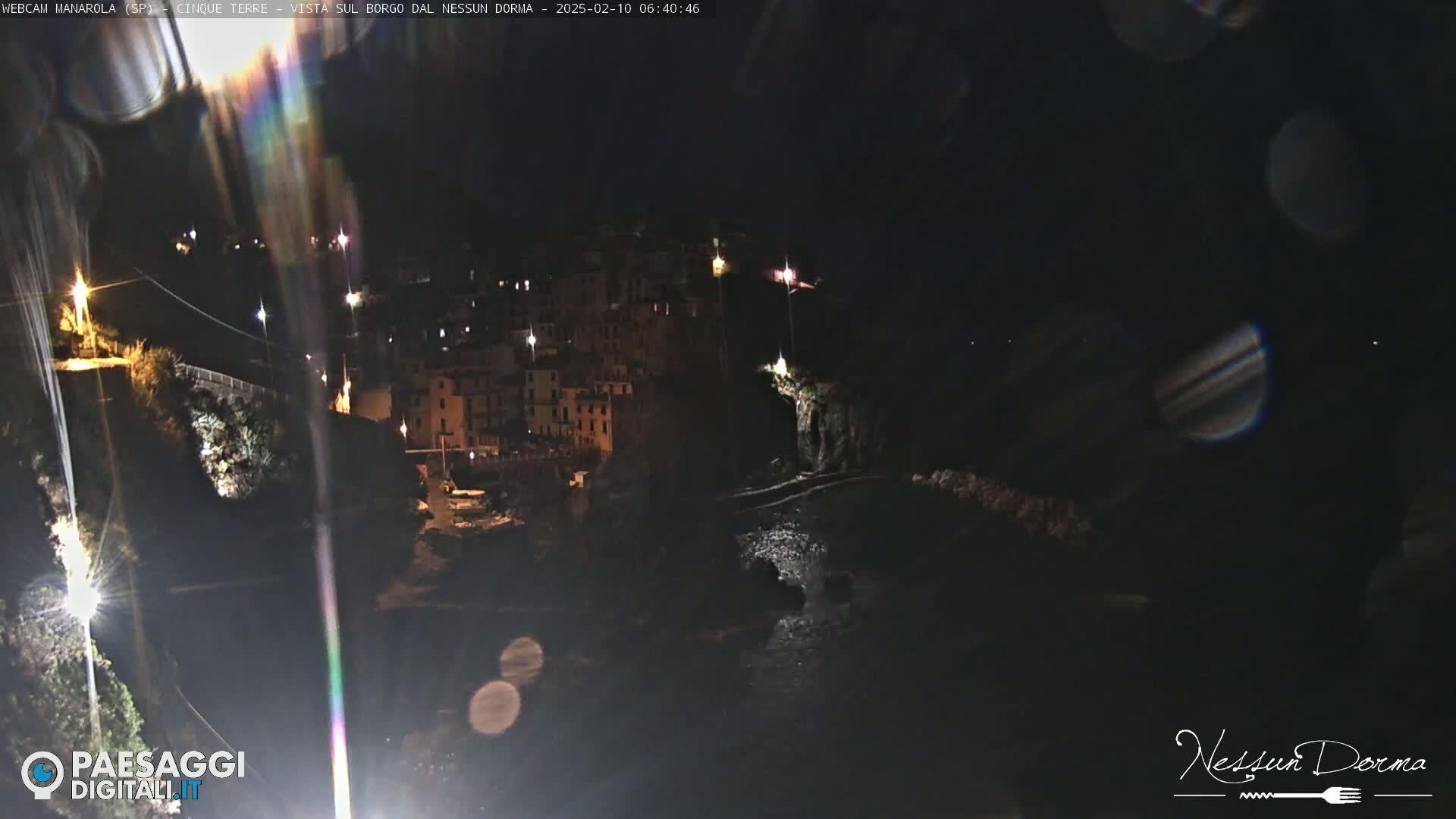 A nighttime view of a coastal town with buildings illuminated by scattered lights, seen from a vantage point above.