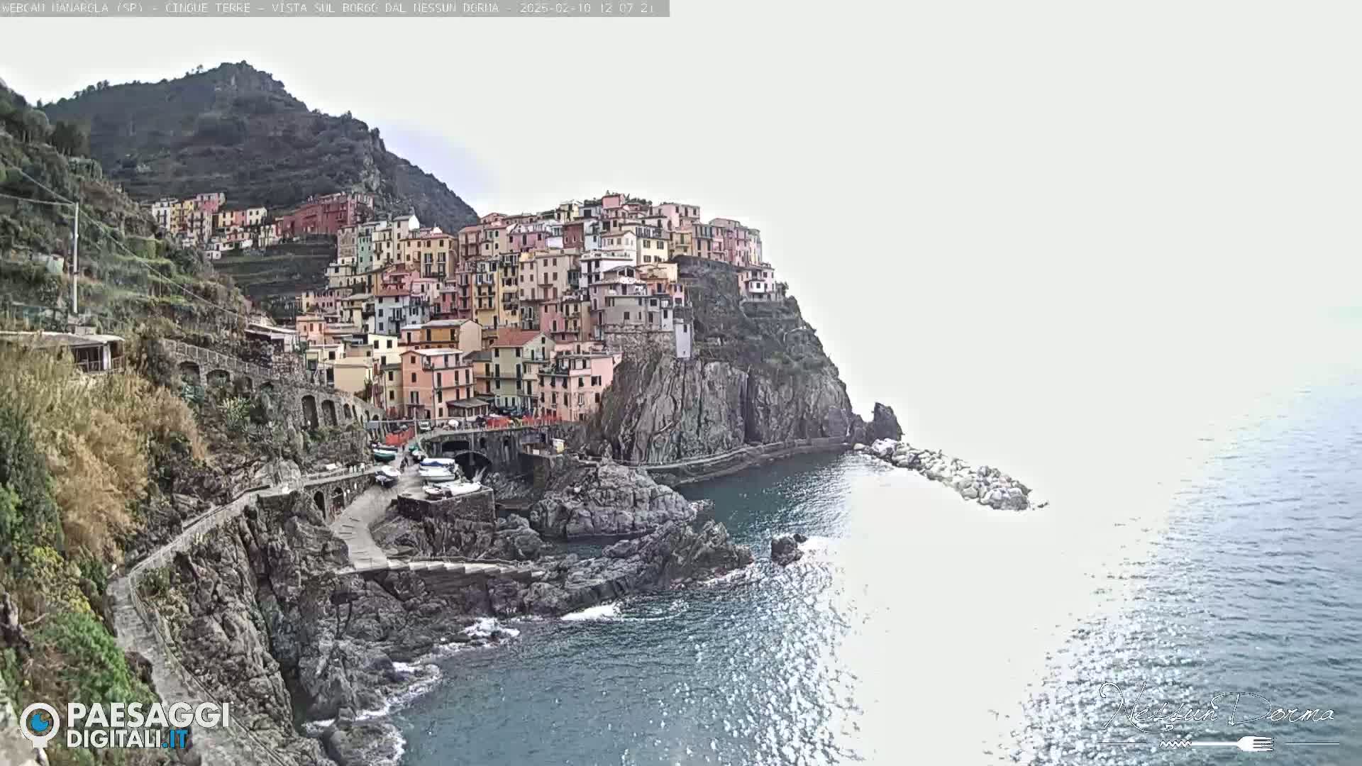 A colorful village clings to a steep cliffside overlooking a calm sea under a mostly cloudy sky.