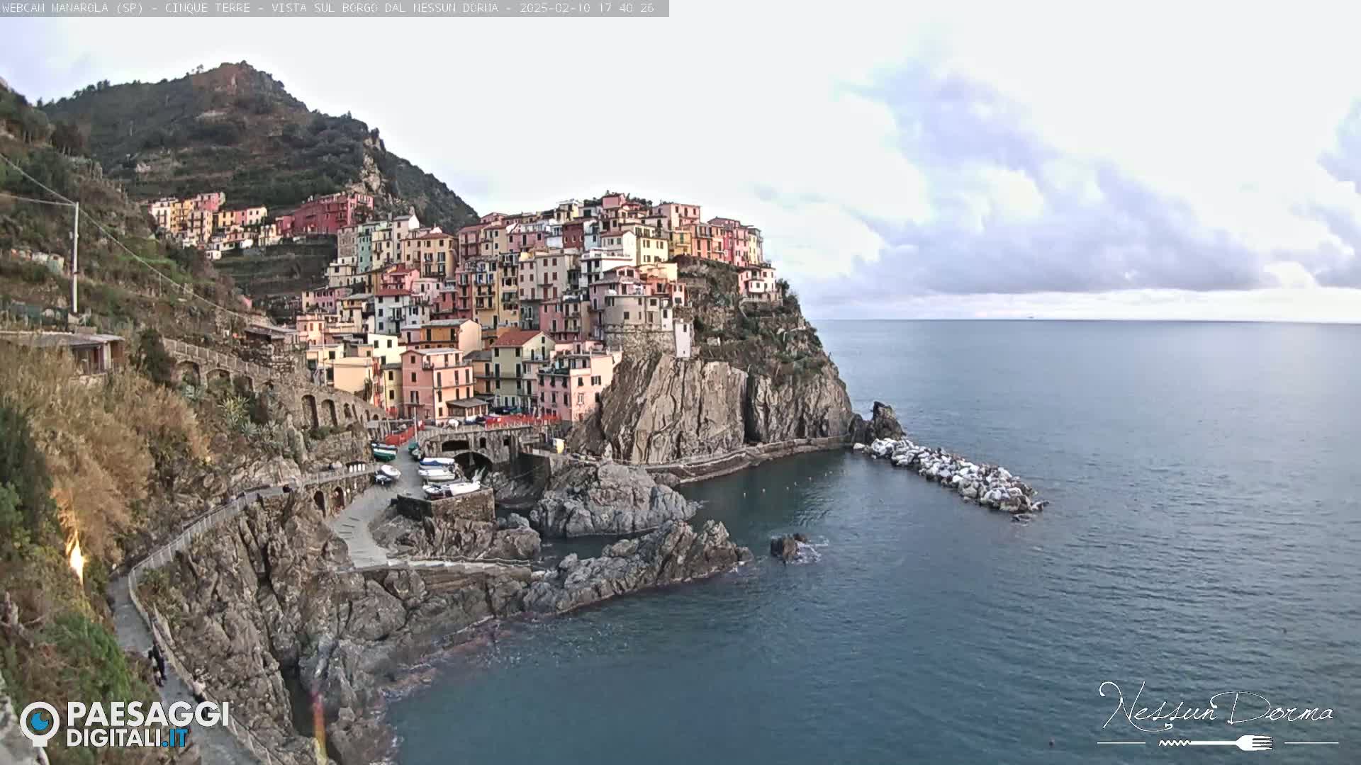A colorful village clings to a steep cliffside overlooking a calm sea under a cloudy sky.