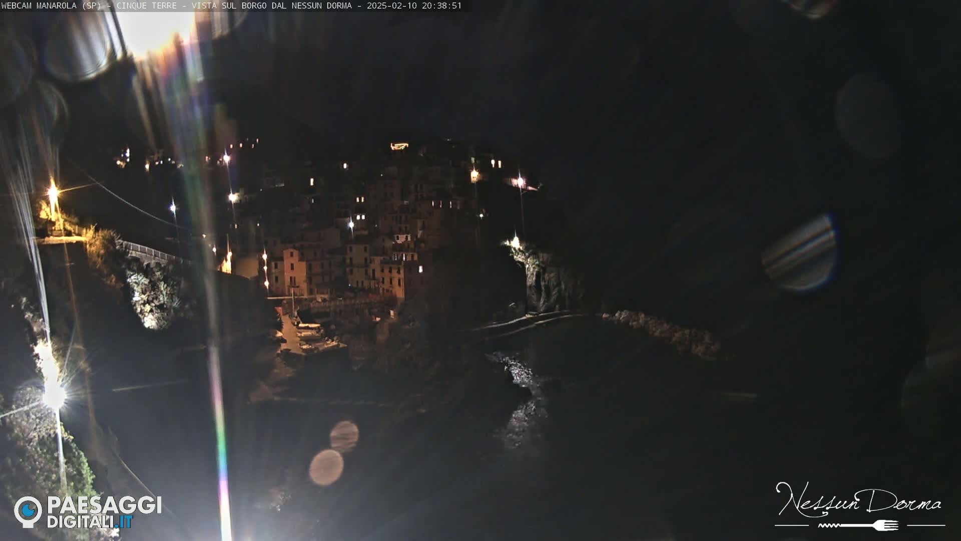 A nighttime view of a coastal town illuminated by lights, with dark water visible below and lens flares present in the image.