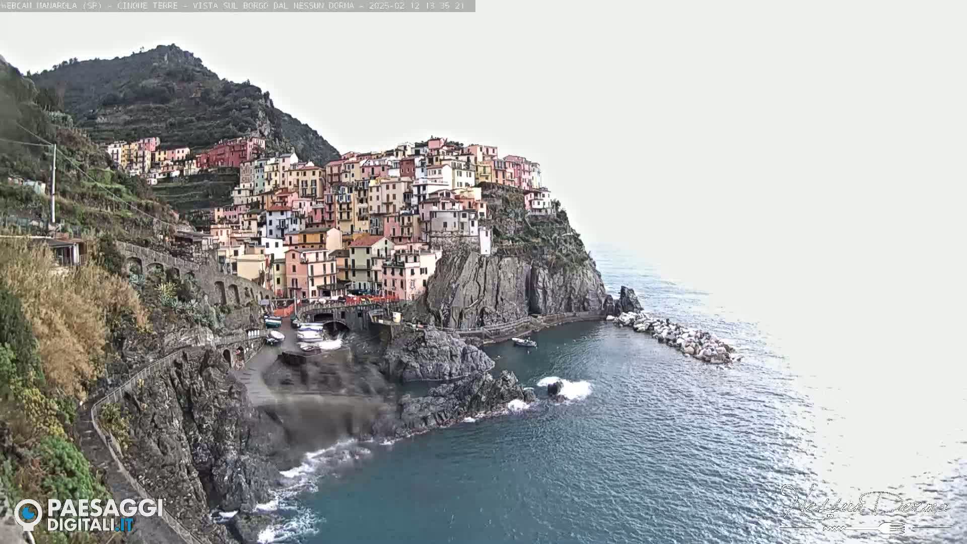 A colorful village clings to a steep cliffside overlooking a choppy sea under an overcast sky.