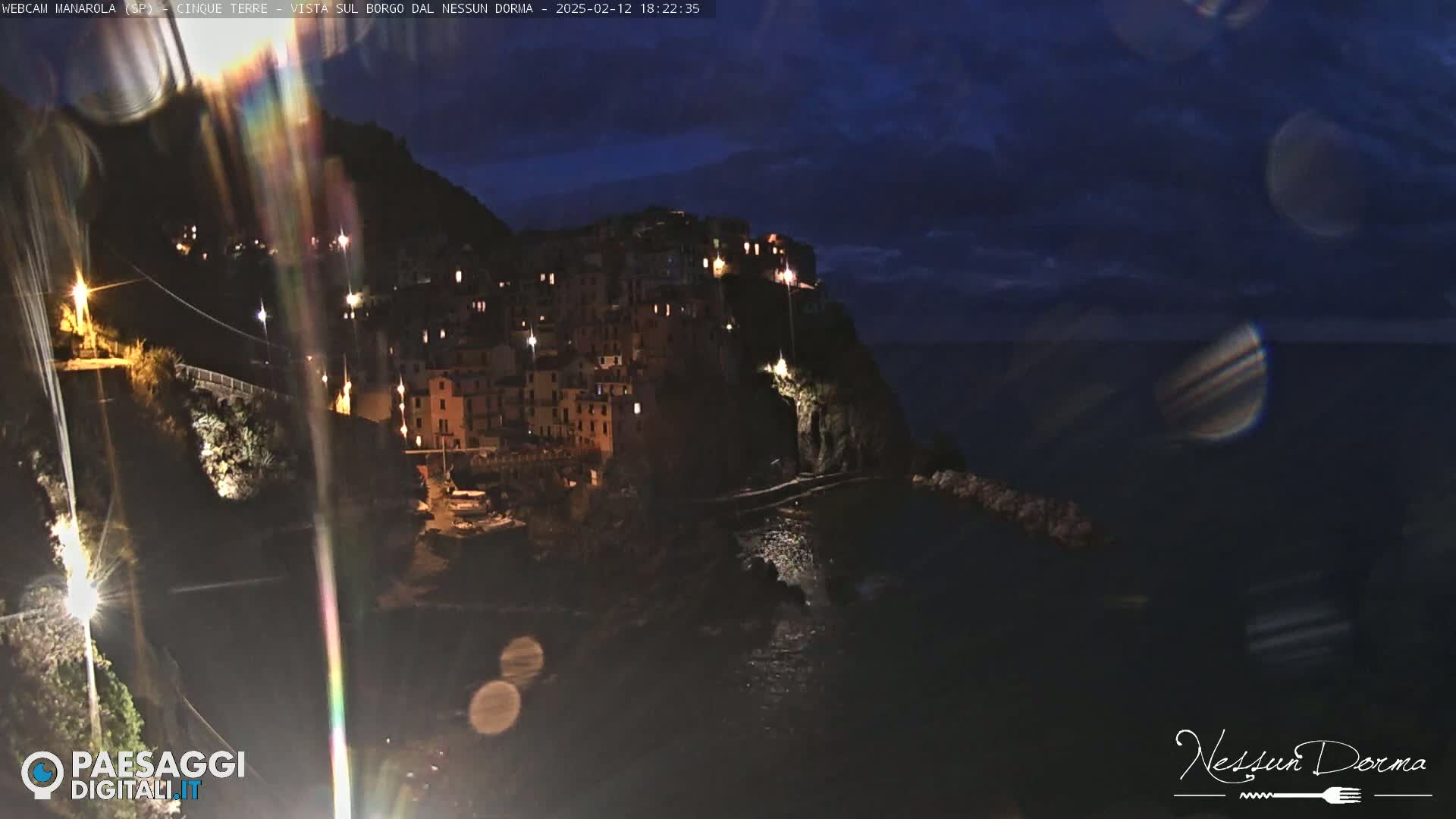 A nighttime coastal village clings to a cliffside, illuminated by scattered lights under a dark, cloudy sky.