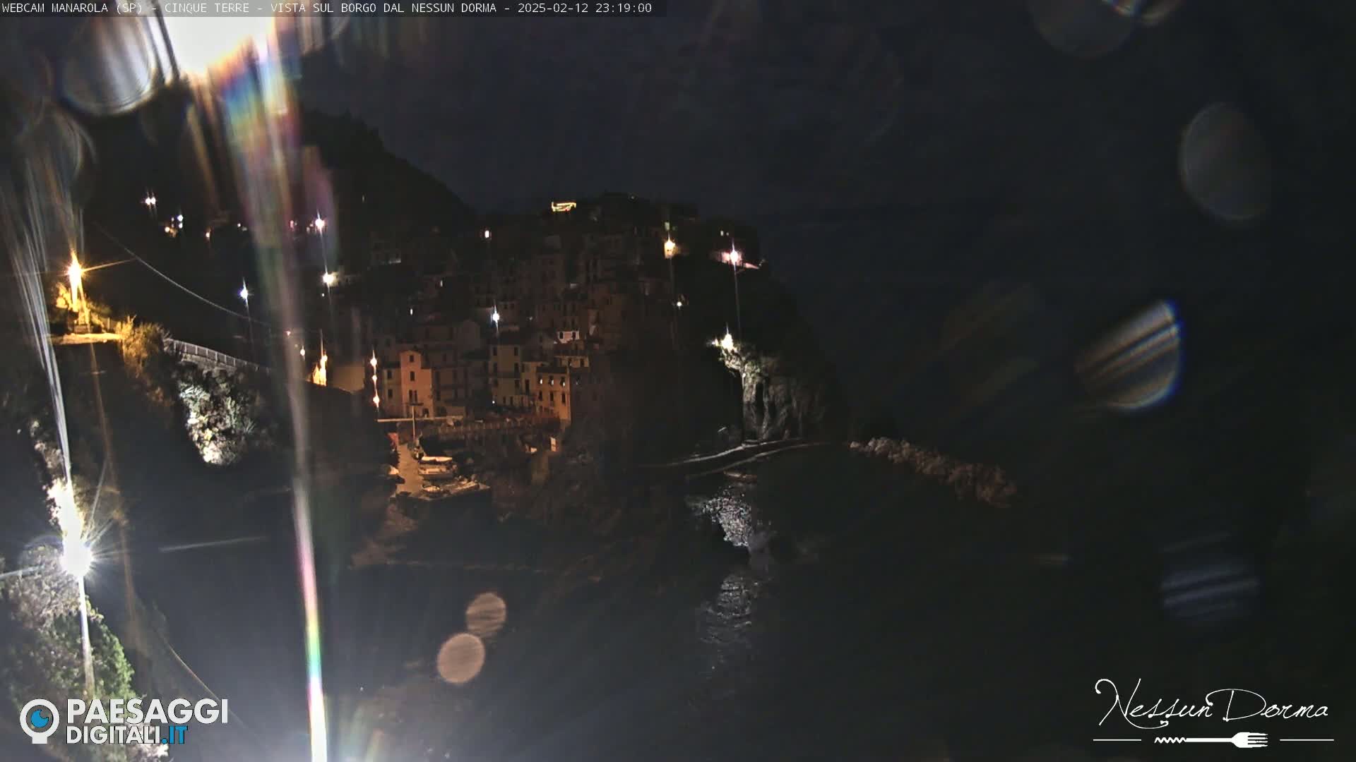 A nighttime view of a coastal town illuminated by scattered lights, with dark water below and lens flares visible in the image.
