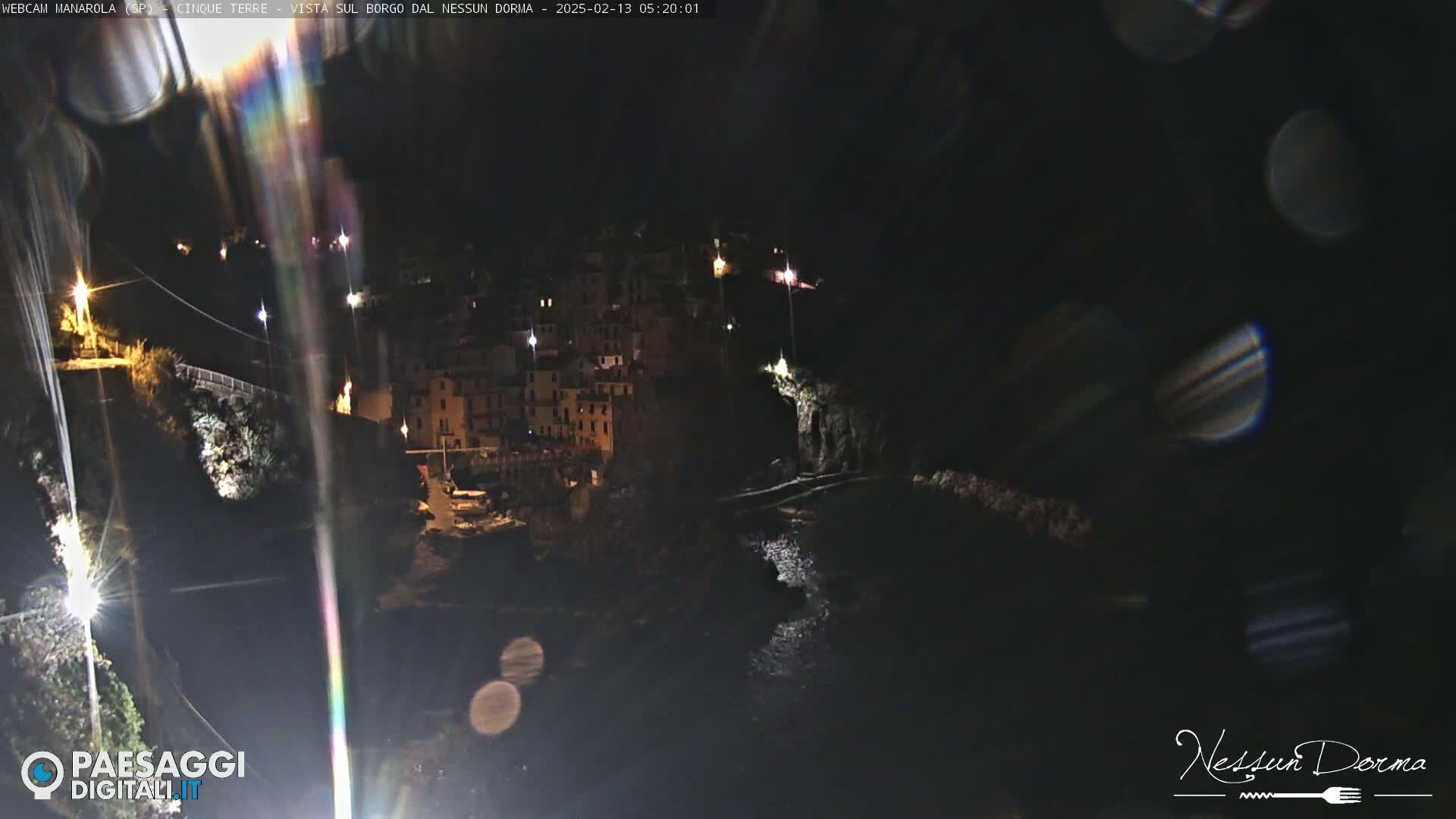 A nighttime view of a coastal town nestled against dark cliffs, illuminated by scattered lights, with a dark body of water visible below.