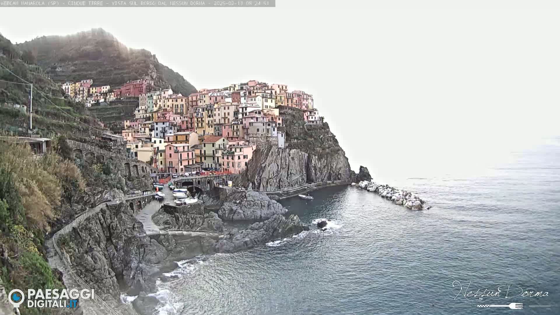 A colorful village clings to a rocky cliffside overlooking a calm sea under a hazy sky.