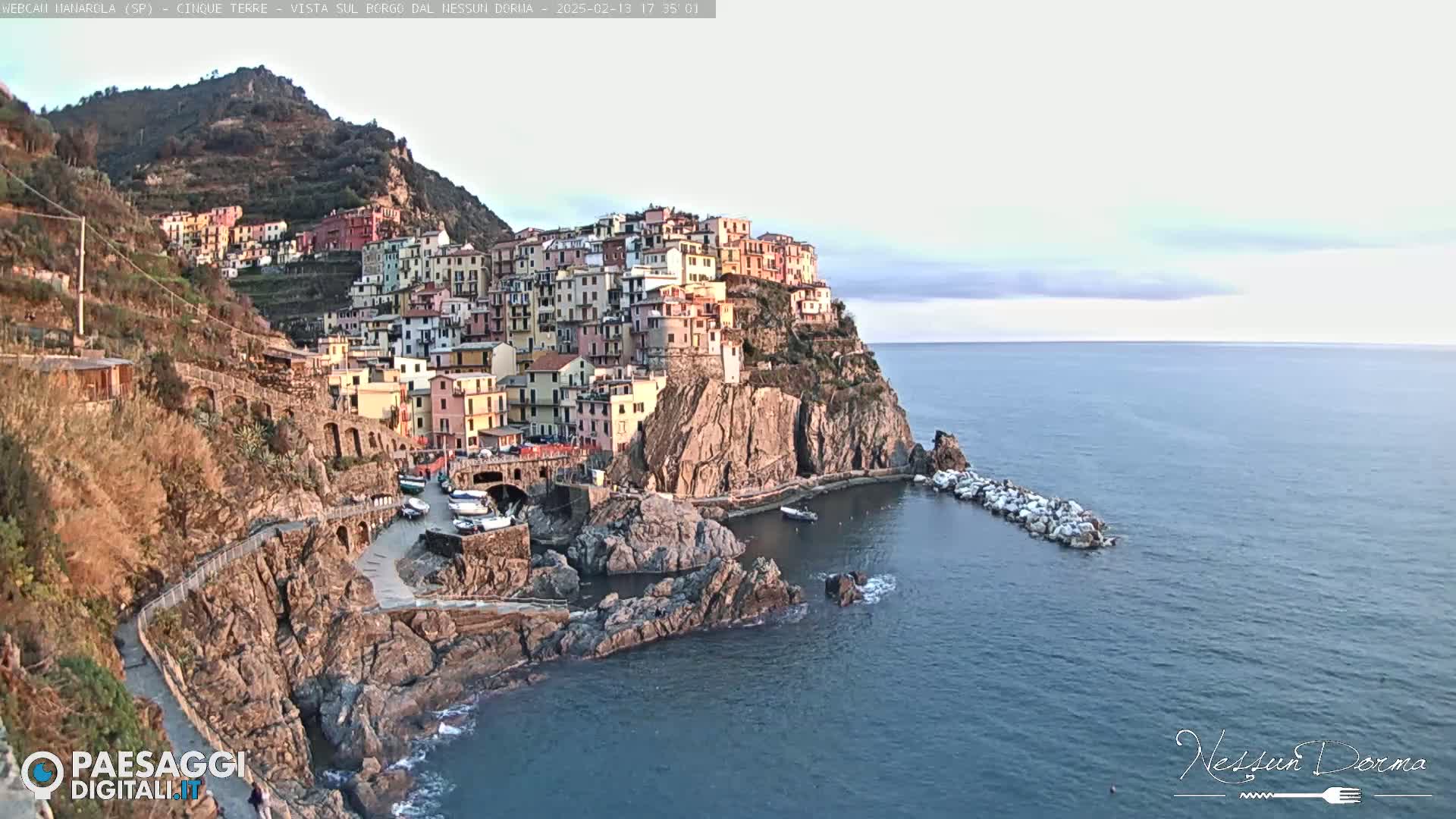 A colorful village clings to a rocky cliffside overlooking a calm sea under a mostly clear sky.