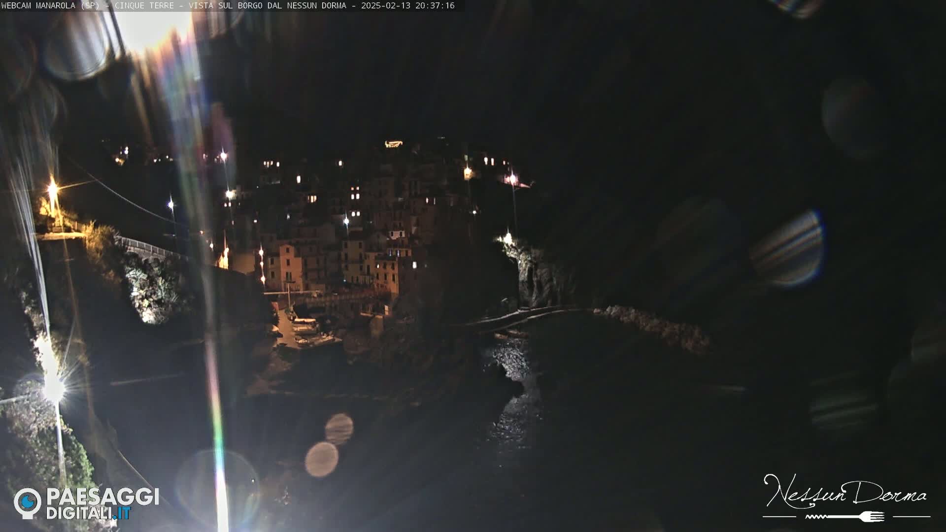 A nighttime view of a coastal town, illuminated by lights, with dark water visible below.