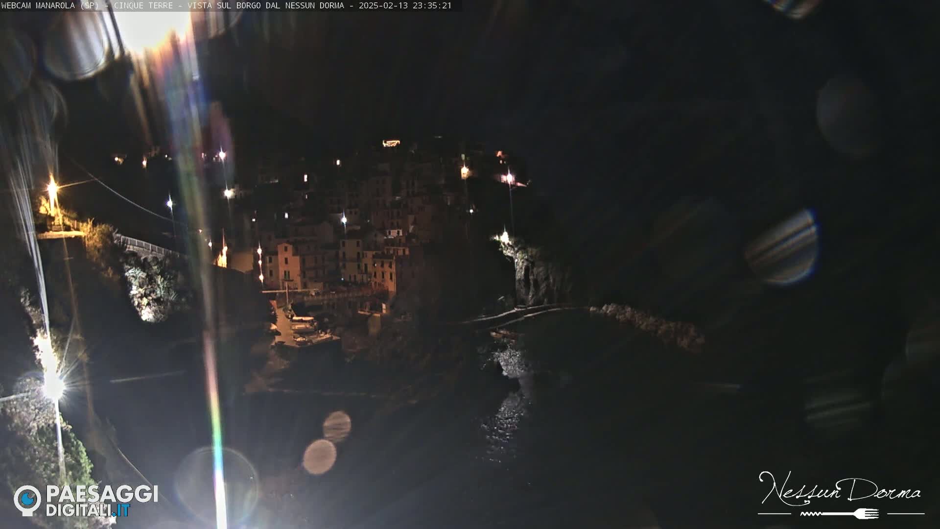 A nighttime view of a coastal town illuminated by lights, with dark water visible below.