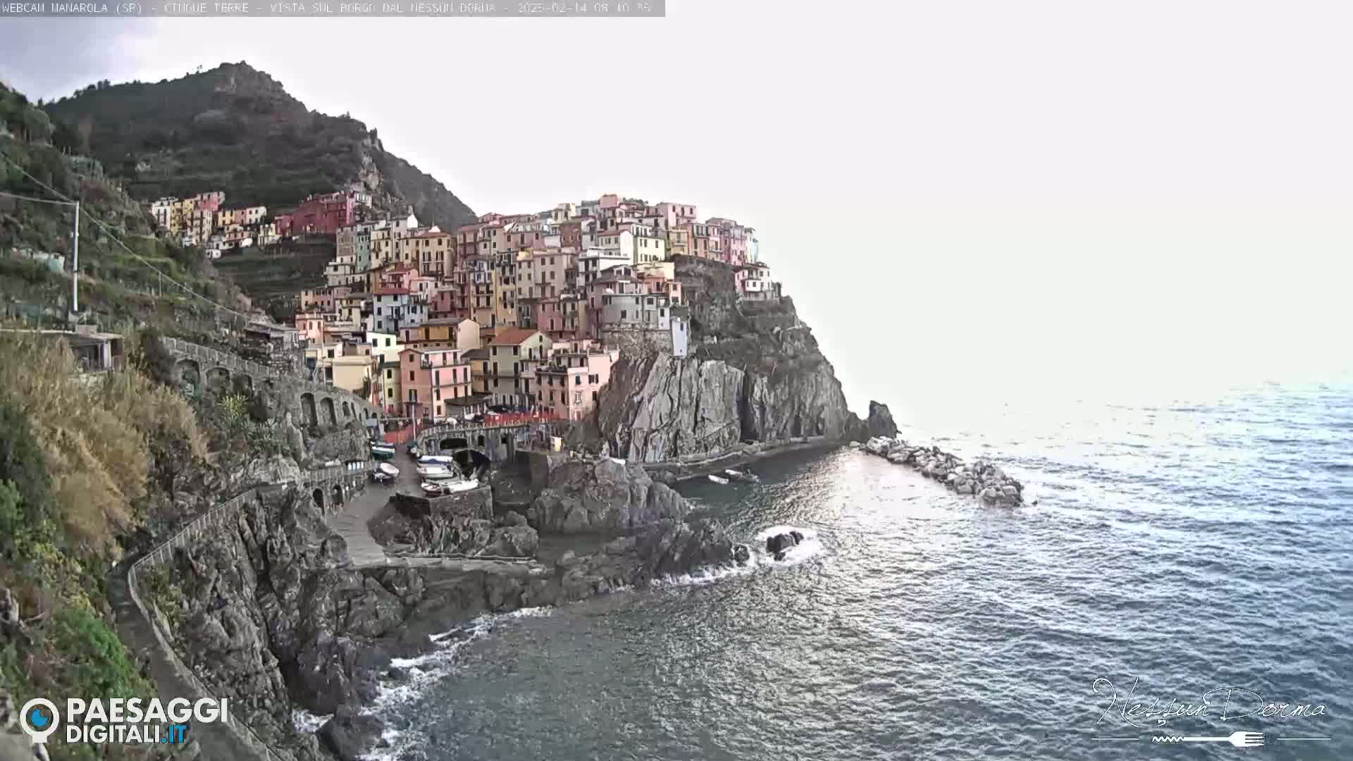 A colorful village clings to a steep cliffside overlooking a calm sea under a bright, overcast sky.
