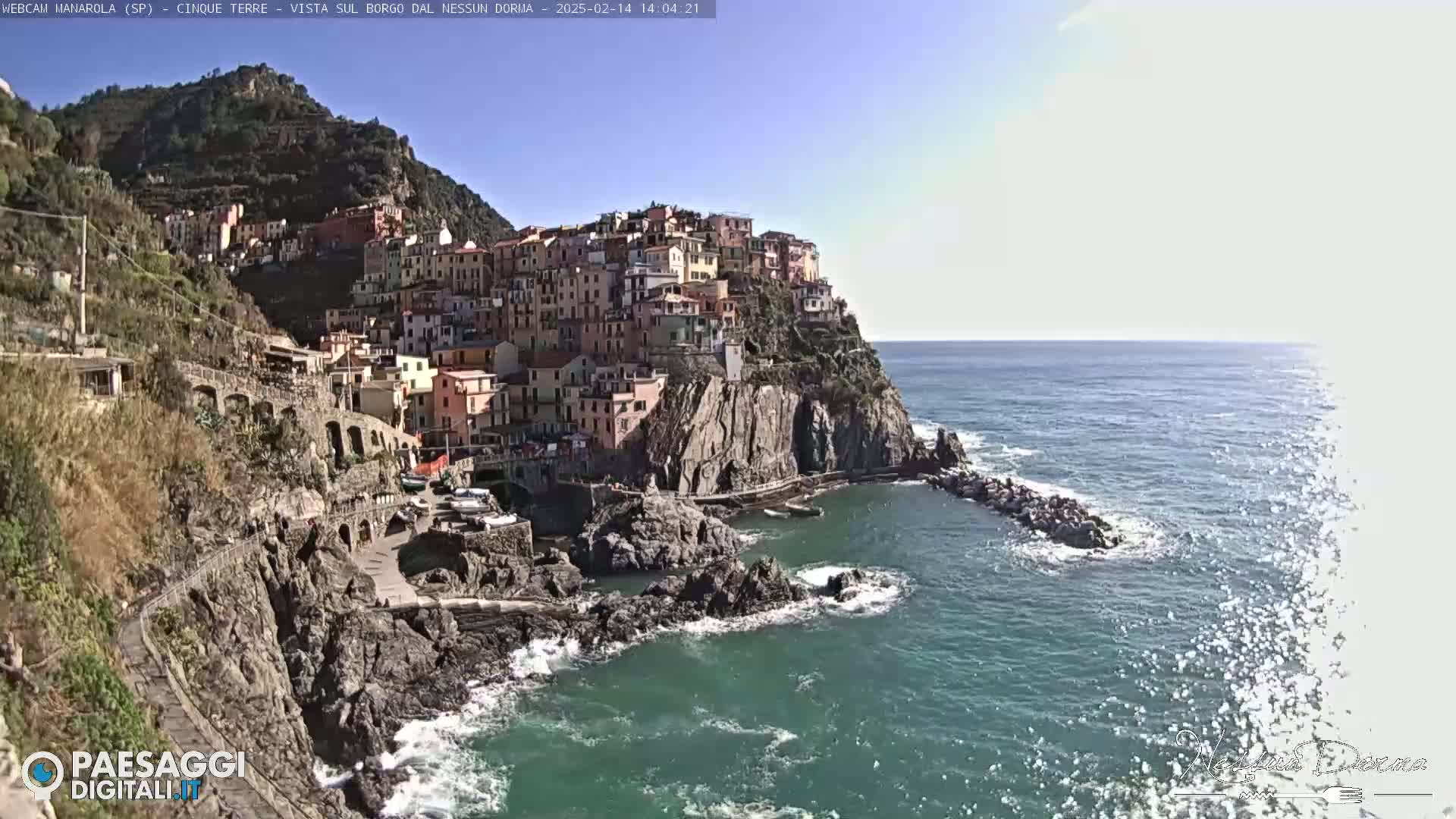 A colorful village clings to a steep cliffside overlooking a blue sea on a sunny day.