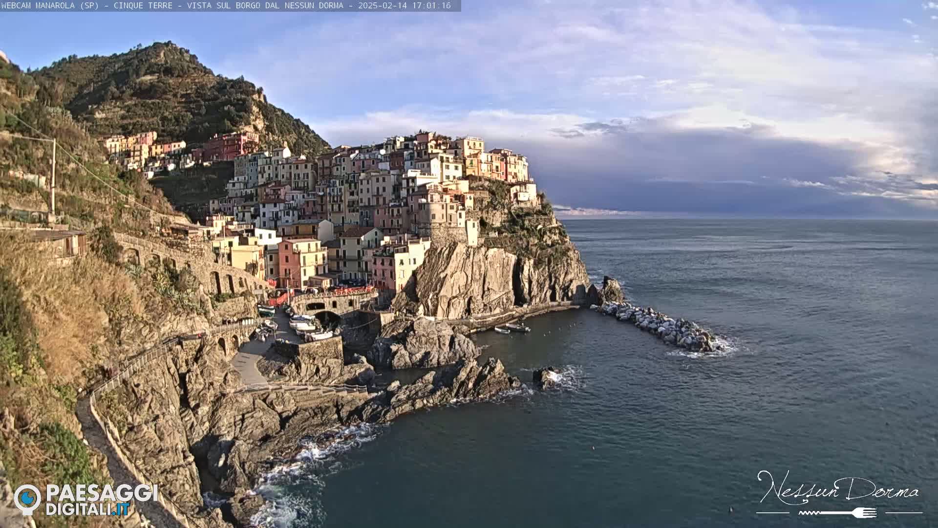 A colorful village clings to a steep cliffside overlooking a calm sea under a partly cloudy sky.